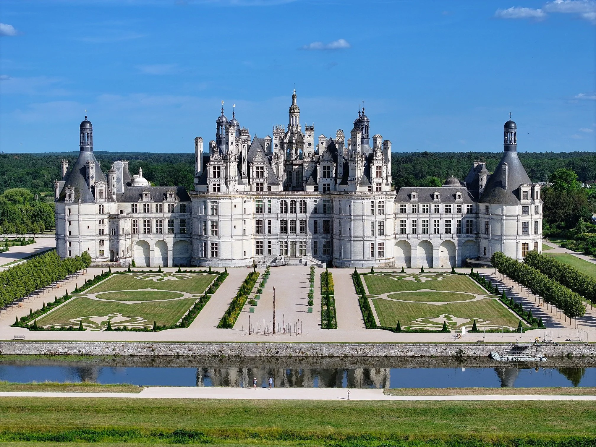 A large white castle with multiple turrets, spires, and ornate architectural details, surrounded by manicured gardens and a water feature in the foreground, with a green forest and blue sky in the background.