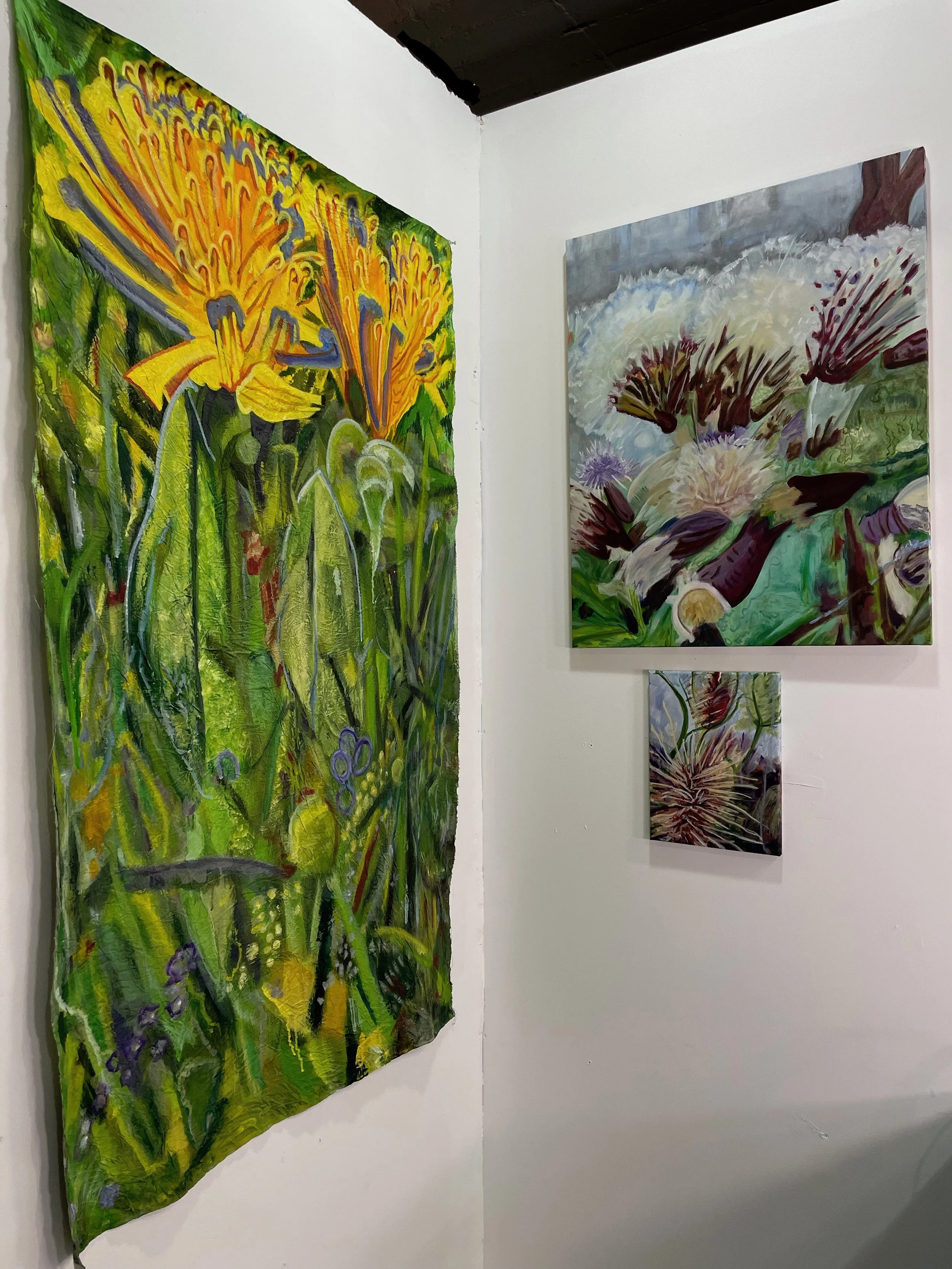 Art gallery wall displaying colorful floral paintings, with a large green and yellow artwork on the left and a smaller purple and white piece on the bottom right.