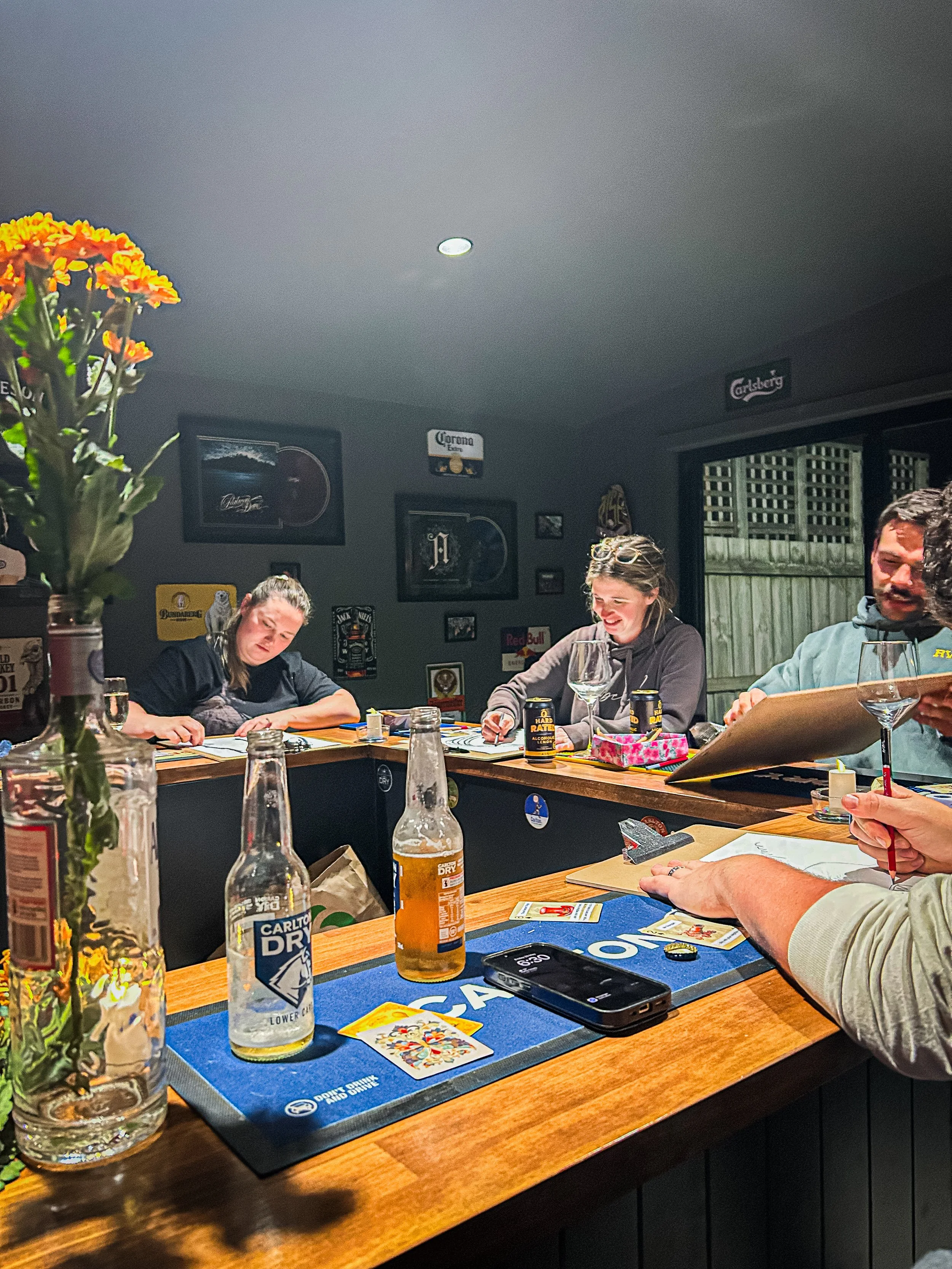 People sitting at a wooden table playing cards in a cozy indoor setting decorated with beer posters and lamps, with a vase of bright orange flowers and bottles of beer on the table.