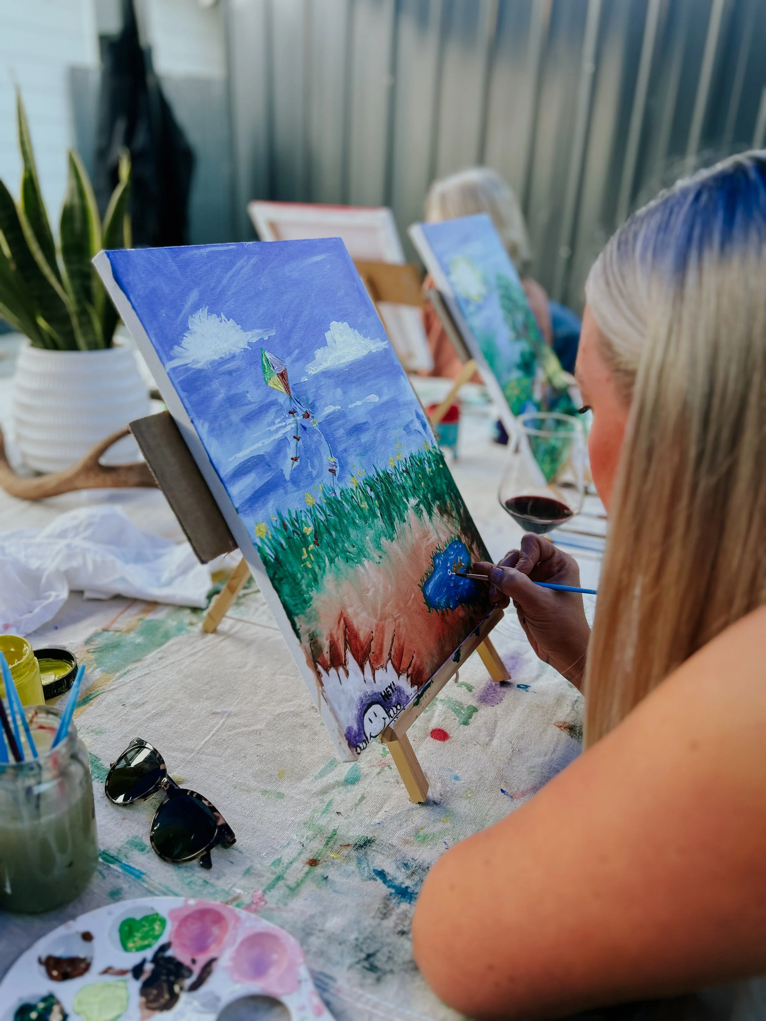 Woman painting at outdoor art class, painting a kite flying in the sky on a canvas, with other people also painting.