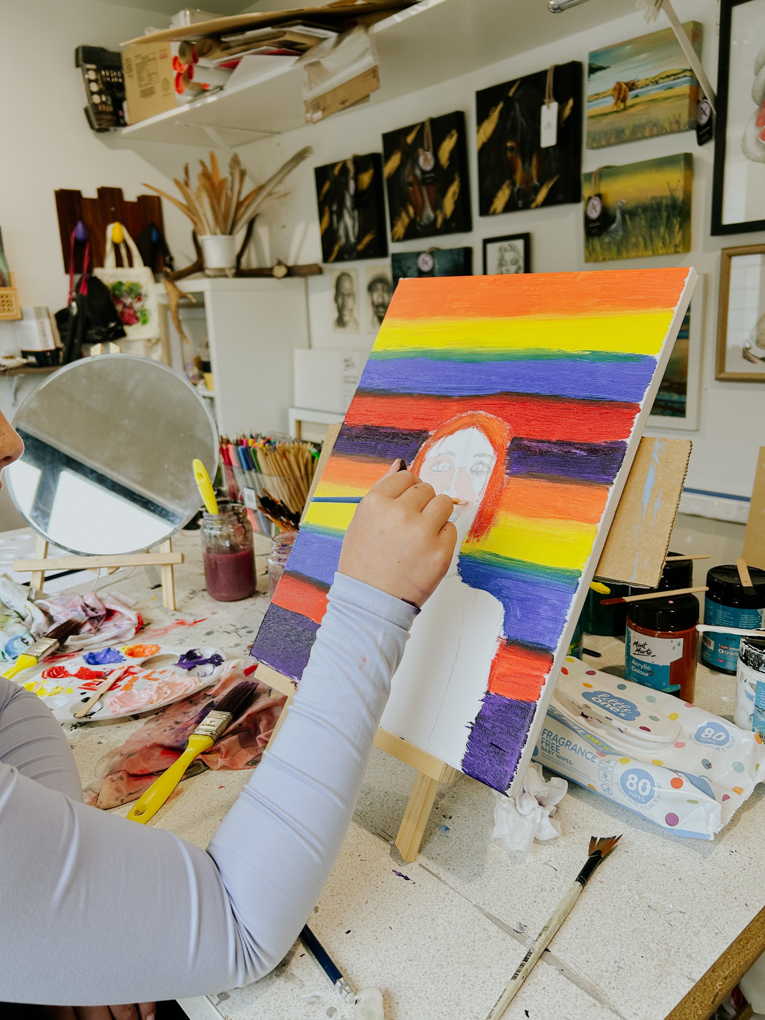 Person painting a portrait of a person's face on a colorful striped background on a canvas in an art studio with various artwork and supplies around.