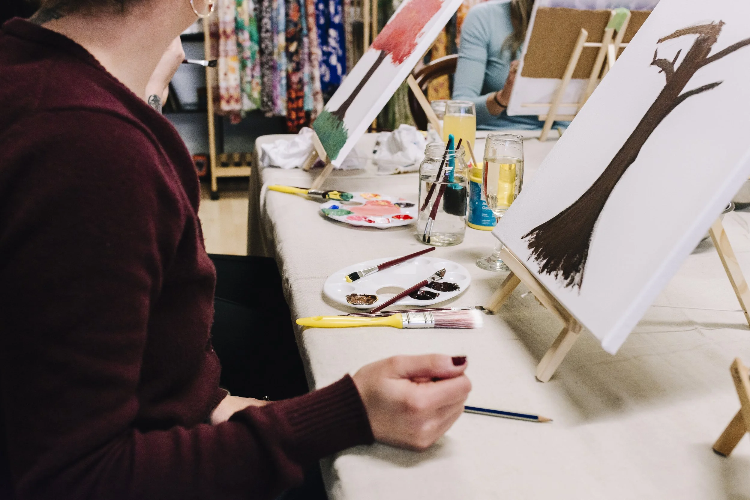 Person painting a tree on canvas at an art class with paints, brushes, and drinks on the table.