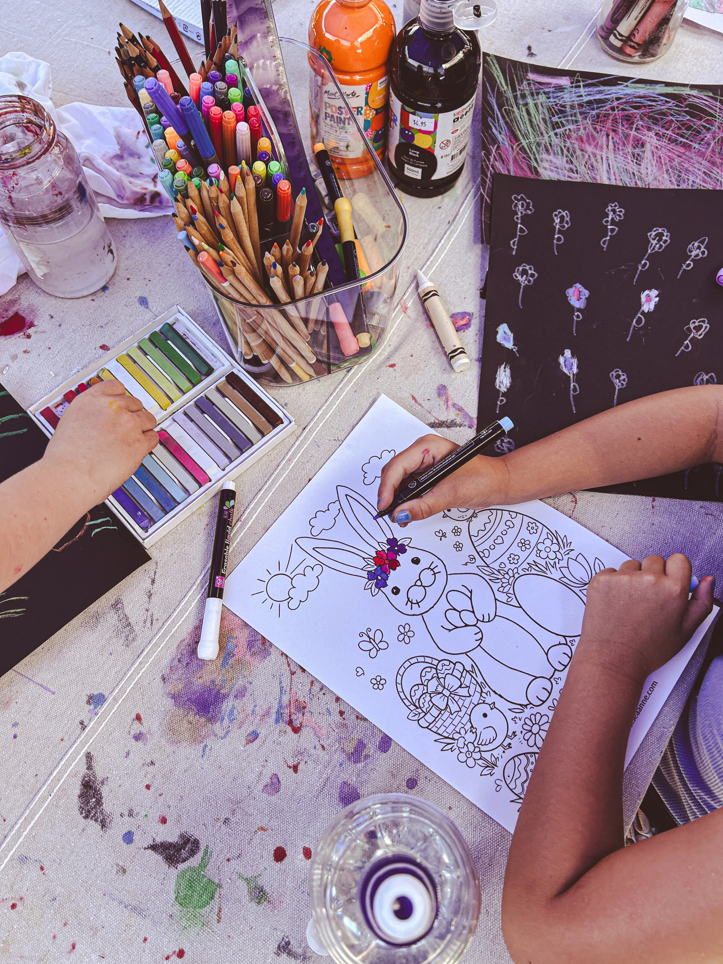 Children coloring a large printout of a bunny with flowers and Easter eggs, surrounded by art supplies including colored pastels, markers, paint bottles, and paper on a table splattered with paint.