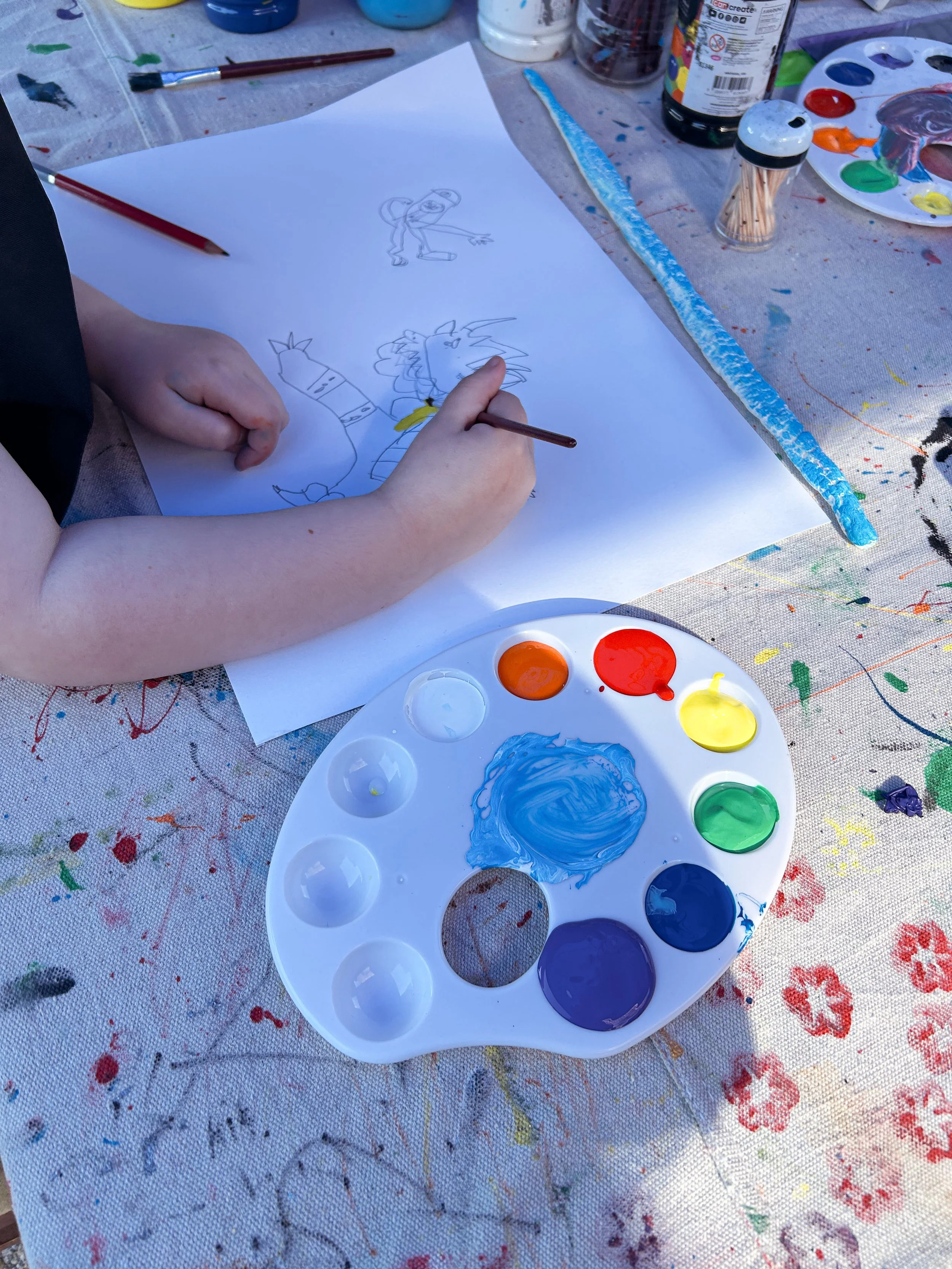 Child drawing cartoon characters on a sheet of paper at a paint table with various art supplies, including a paint palette with blue, yellow, green, red, purple, and orange paints, and a paintbrush.