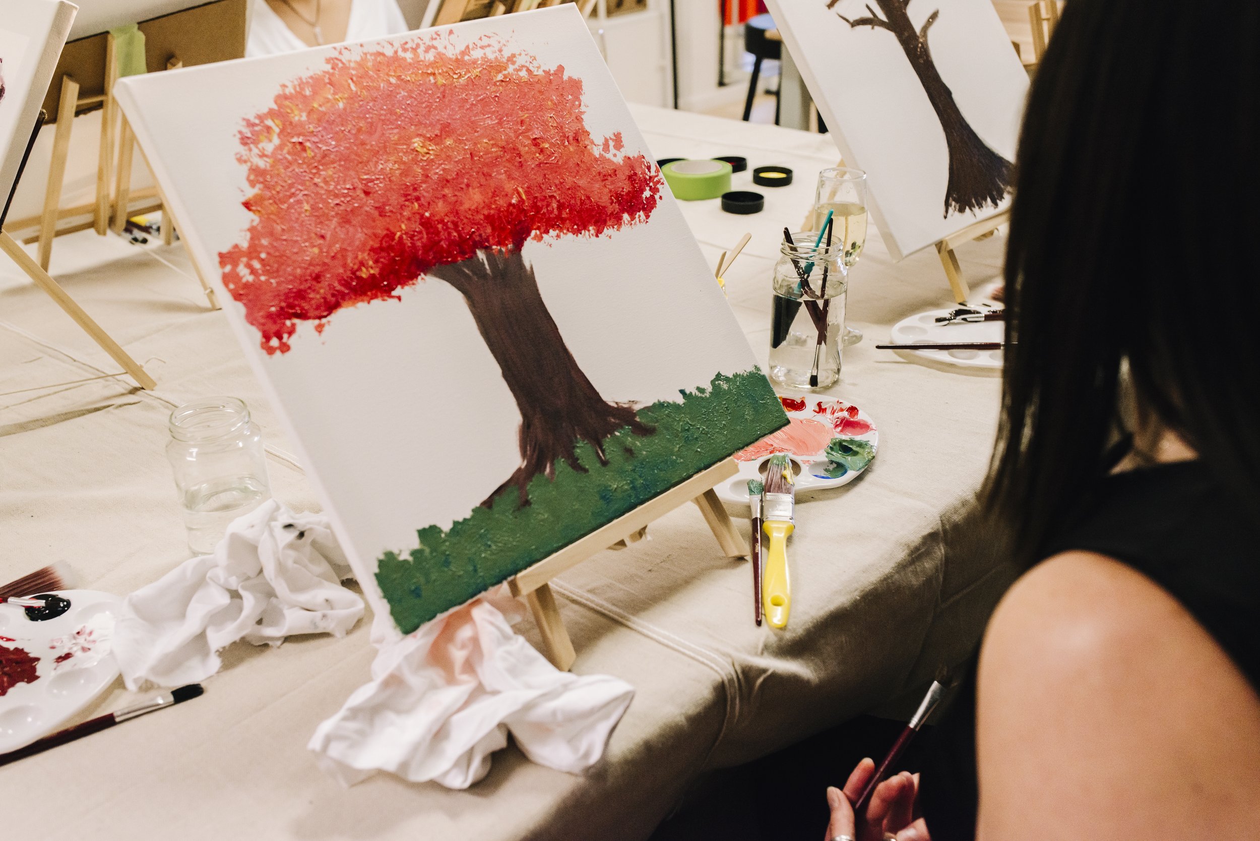 A woman painting a landscape of a tree with red leaves, a brown trunk, and green grass in the background, on a canvas at a painting event.