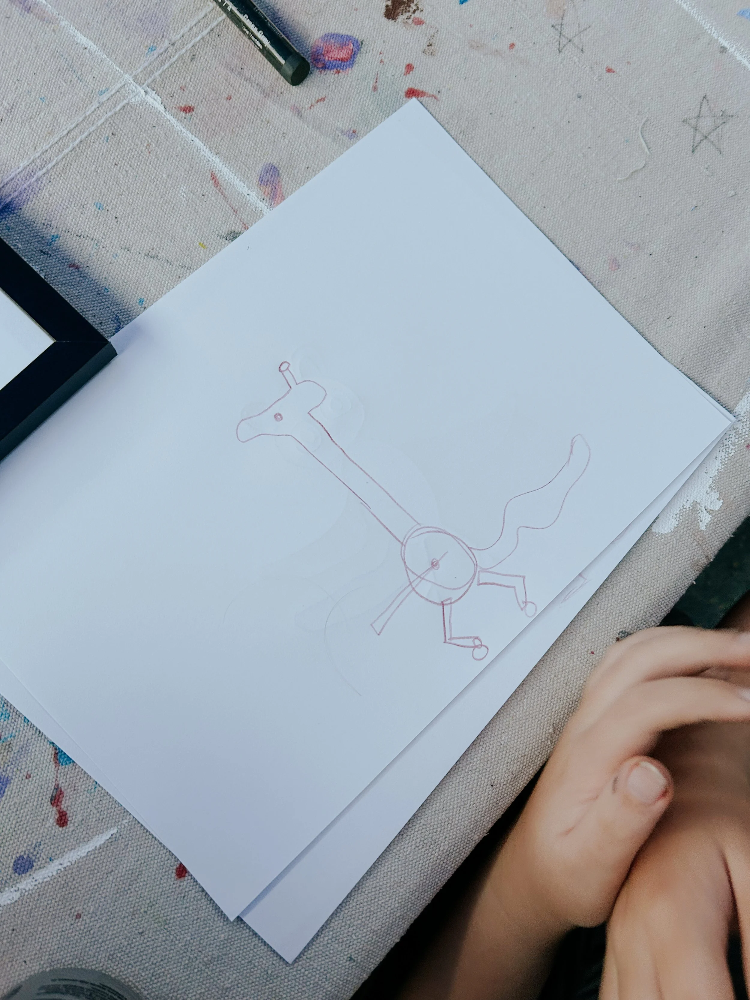 A child's hand rests on a table, with a drawing of an animal mixed up on white paper. 