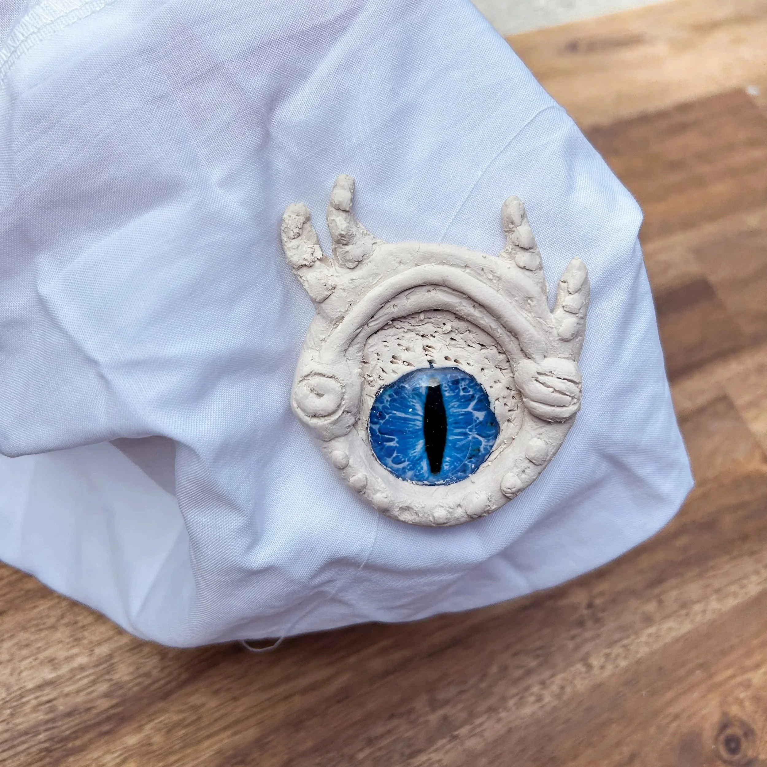 A handmade clay creation resembling a mythical creature with a single large blue eye, white textured body, and horn-like protrusions on top, placed on a white cloth on a wooden surface.