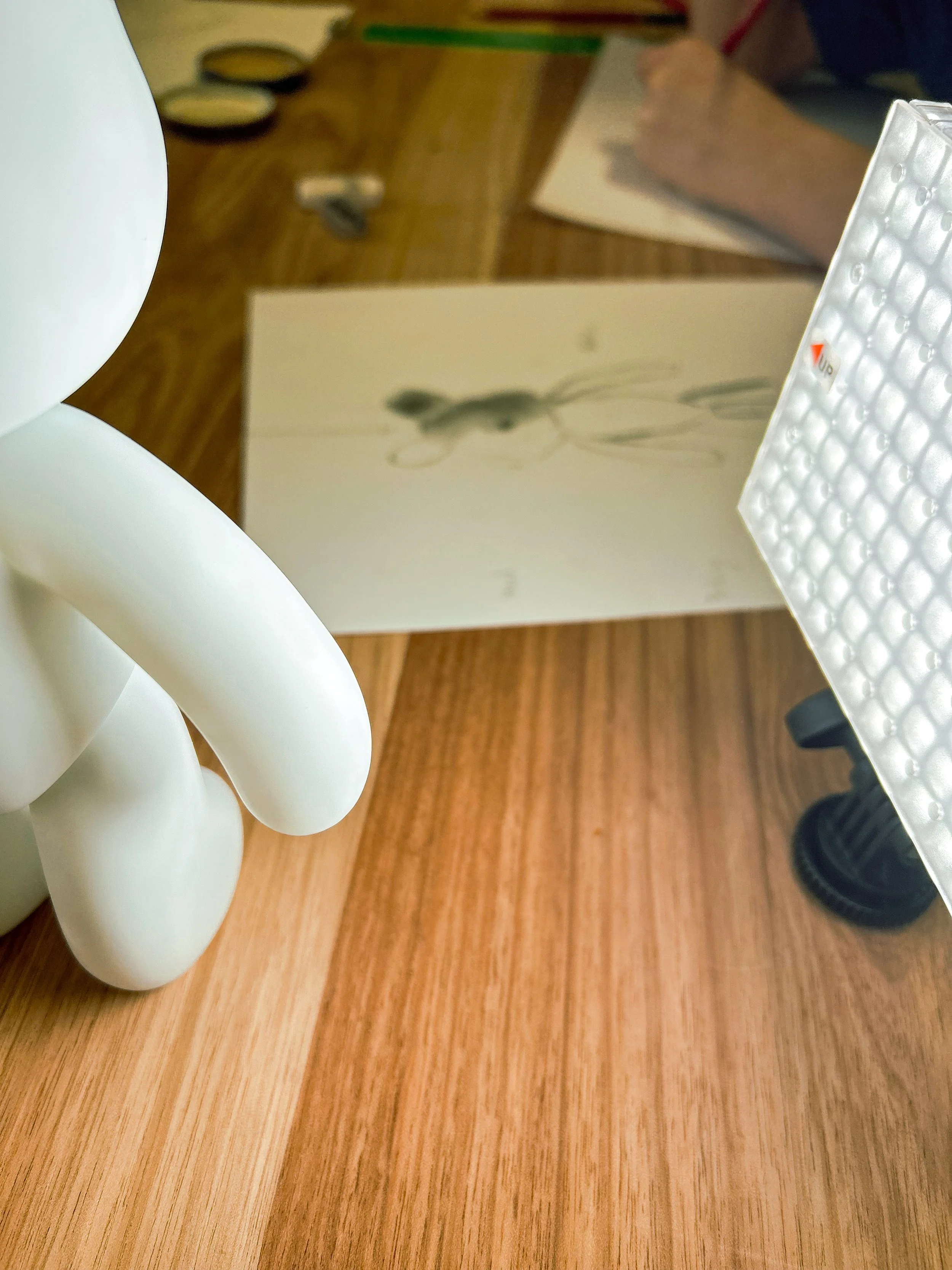 Close-up view of a white sculptural object with rounded, intertwined shapes, a wooden surface, a white textured surface, and a blurred drawing of a woman in the background.