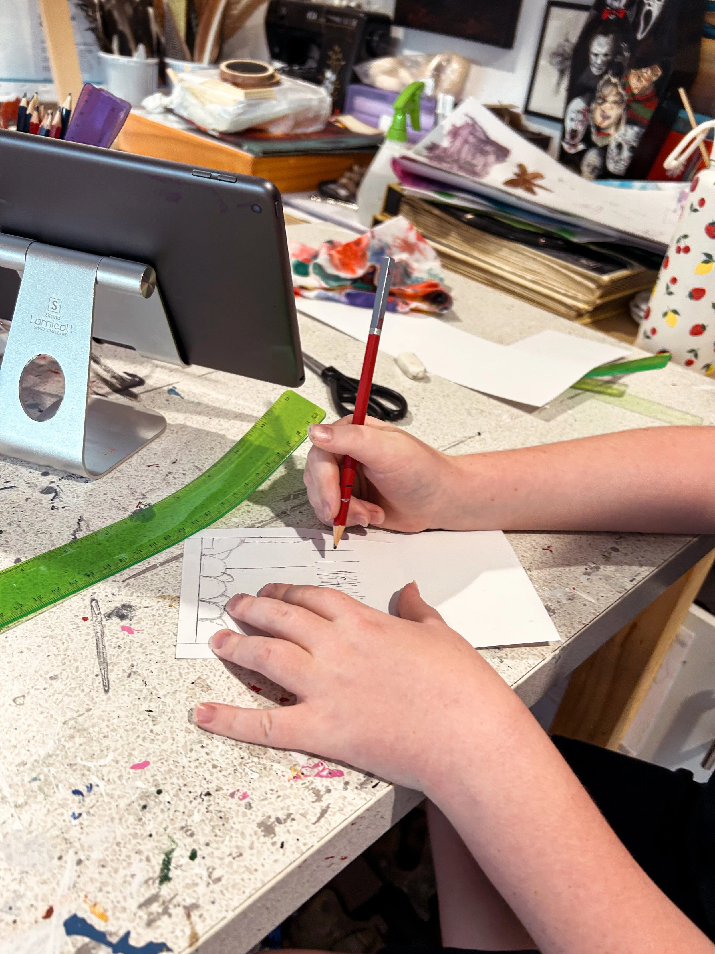 Person's hands drawing a design on a small piece of paper with a red pencil at an art and craft workspace filled with art supplies and sketches.