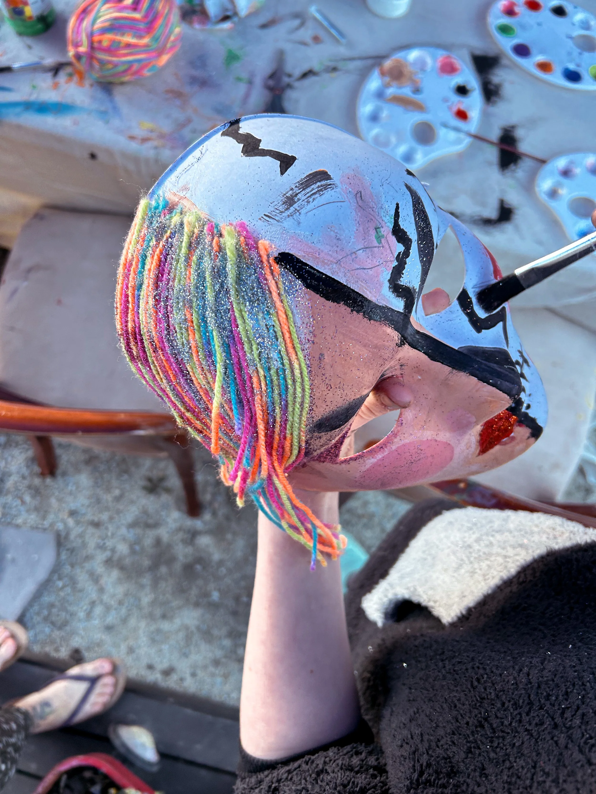 A person holding a decorated ceramic mask with colorful yarn and painted designs at an outdoor art event.