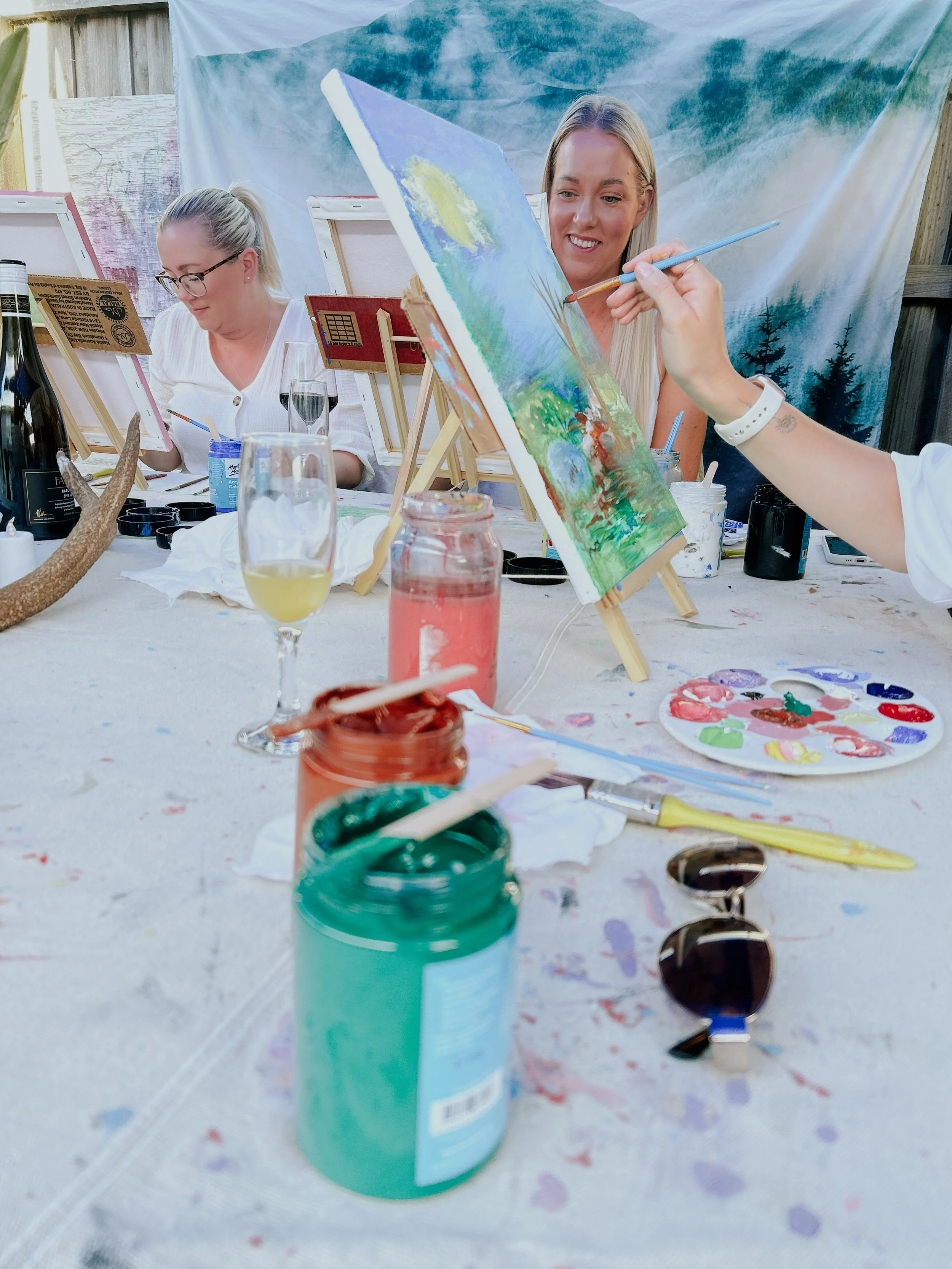 People painting outdoors at a communal art event, with painting supplies and drinks on the table.