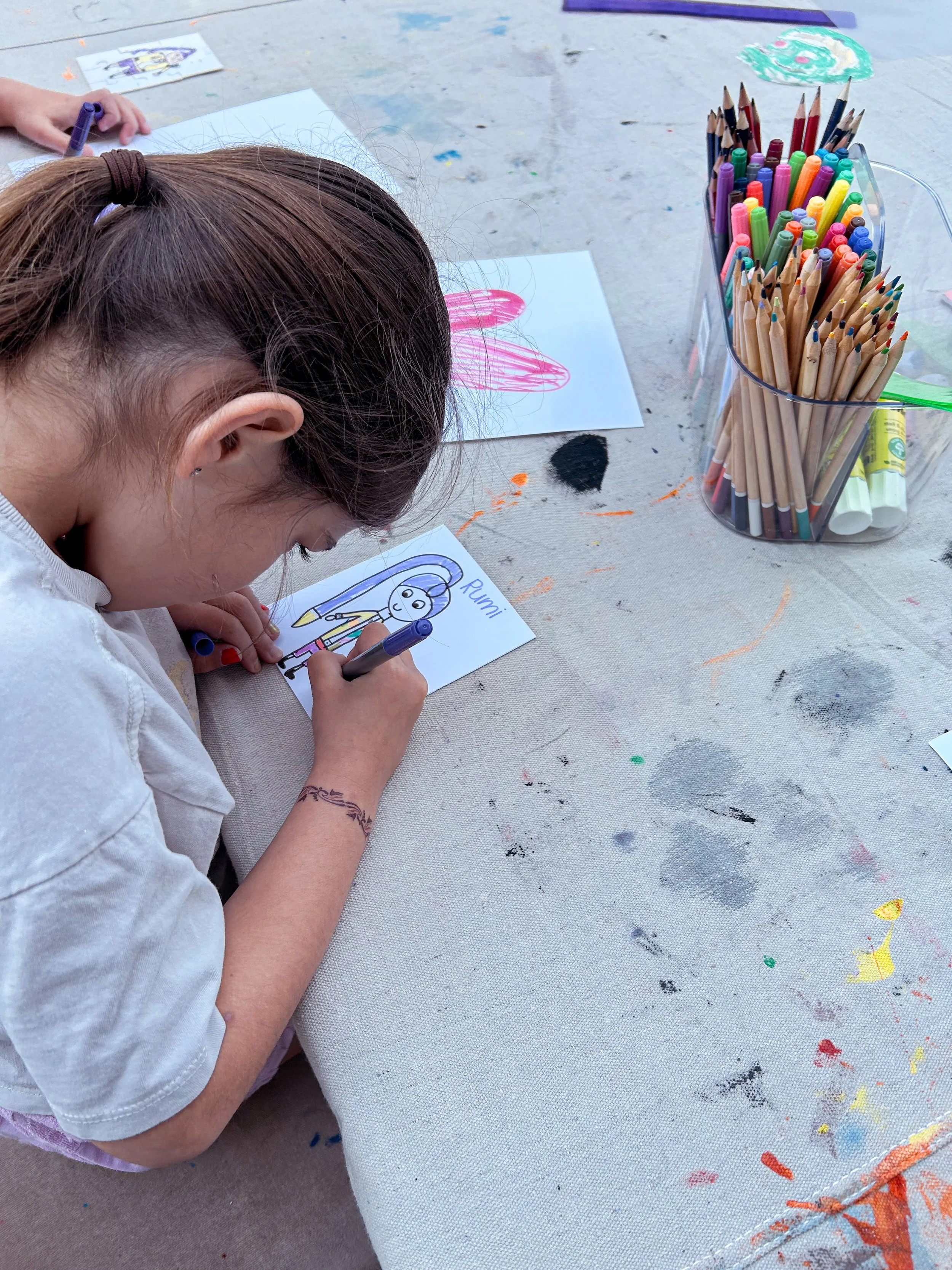 A young girl with brown hair in a ponytail drawing a colorful picture on a small paper using a blue marker, with a container of colored markers and pencils nearby on a paint-stained table.