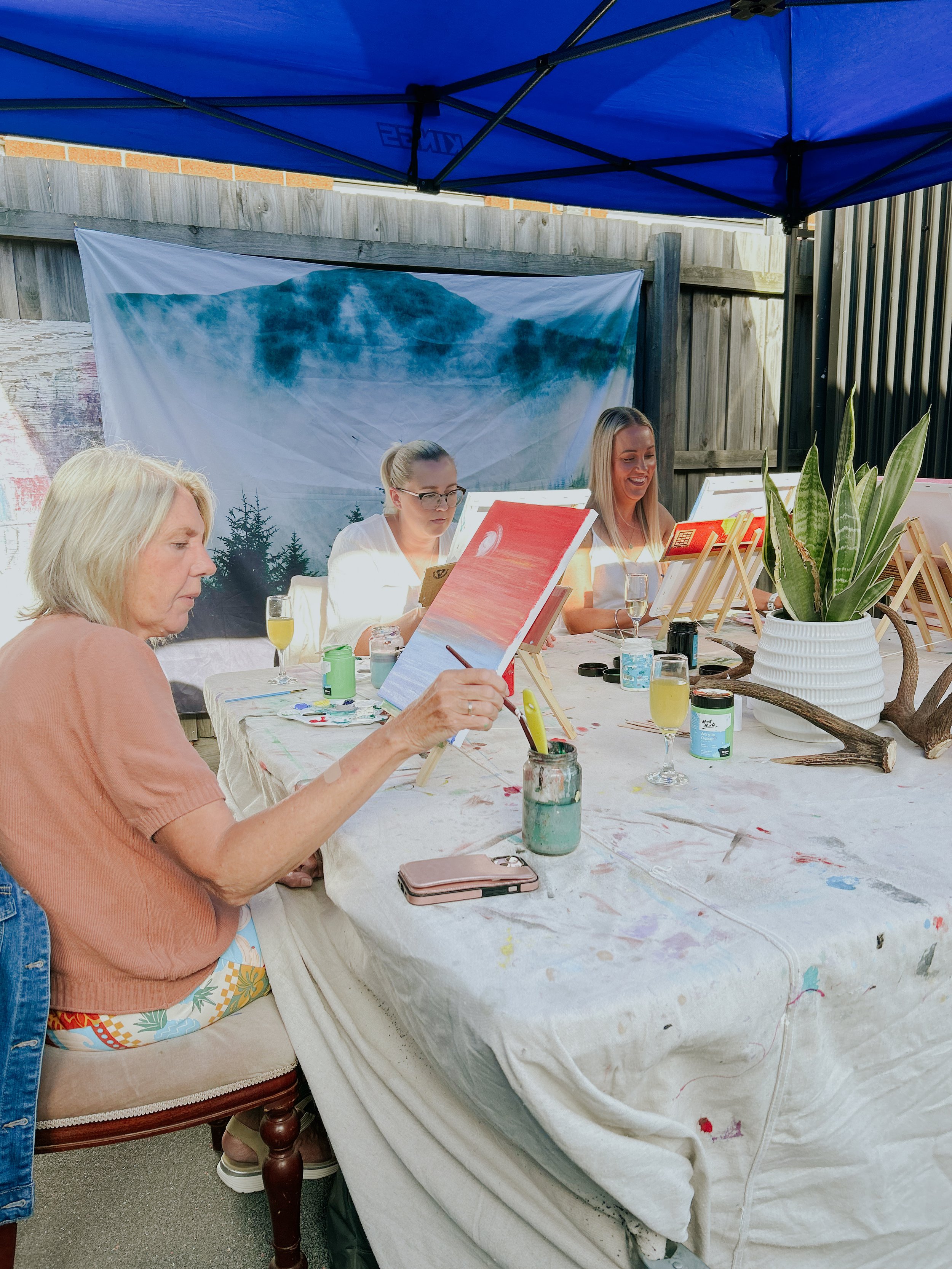 Women painting on canvas at an outdoor art party under a blue canopy in a backyard.