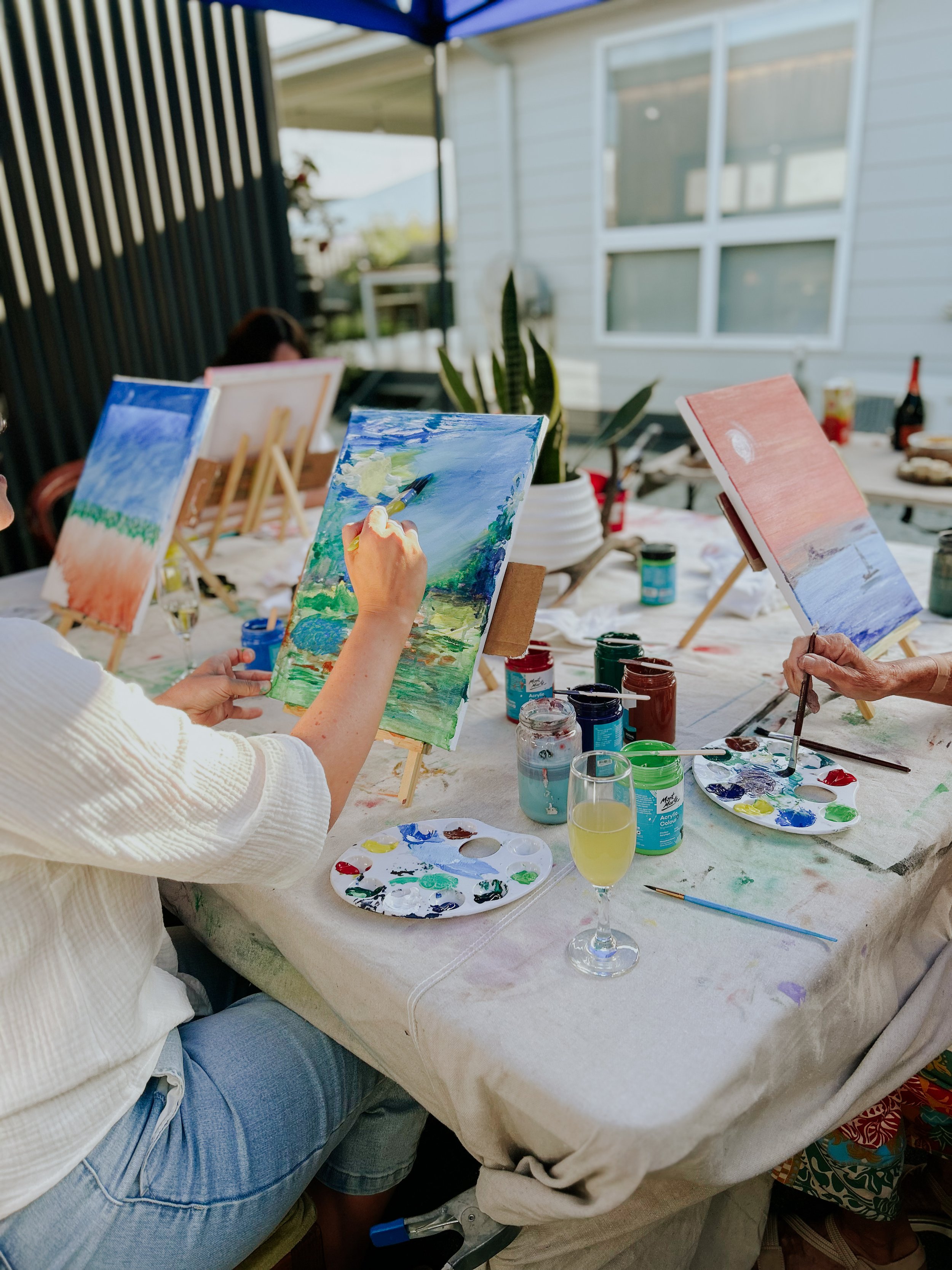 People painting landscape scenes outdoors, with easels, paint palettes, and brushes on a table, under a canopy.