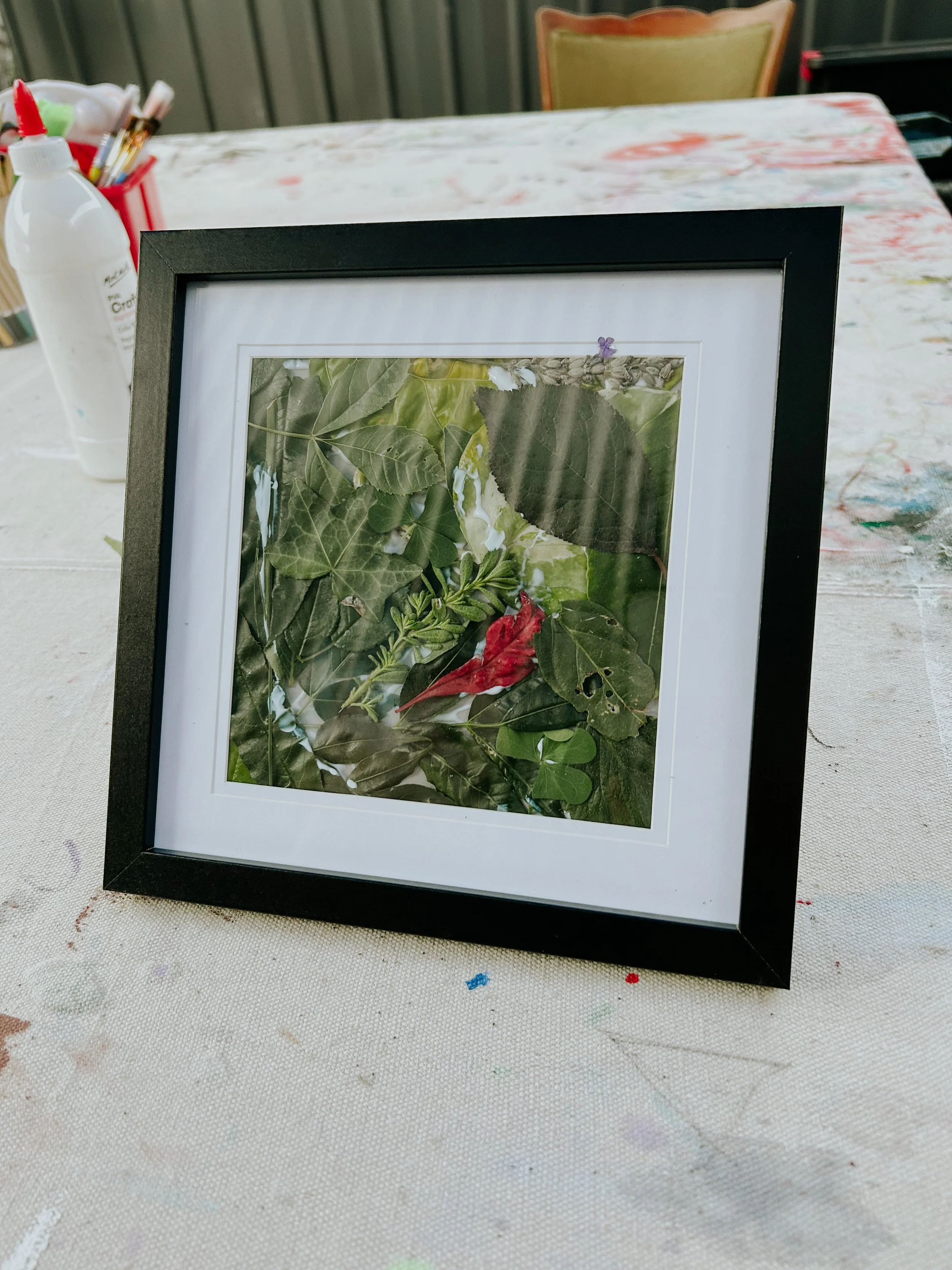 A framed piece of living collage artwork featuring green and red leaves, placed on a table, with art supplies visible in the background.