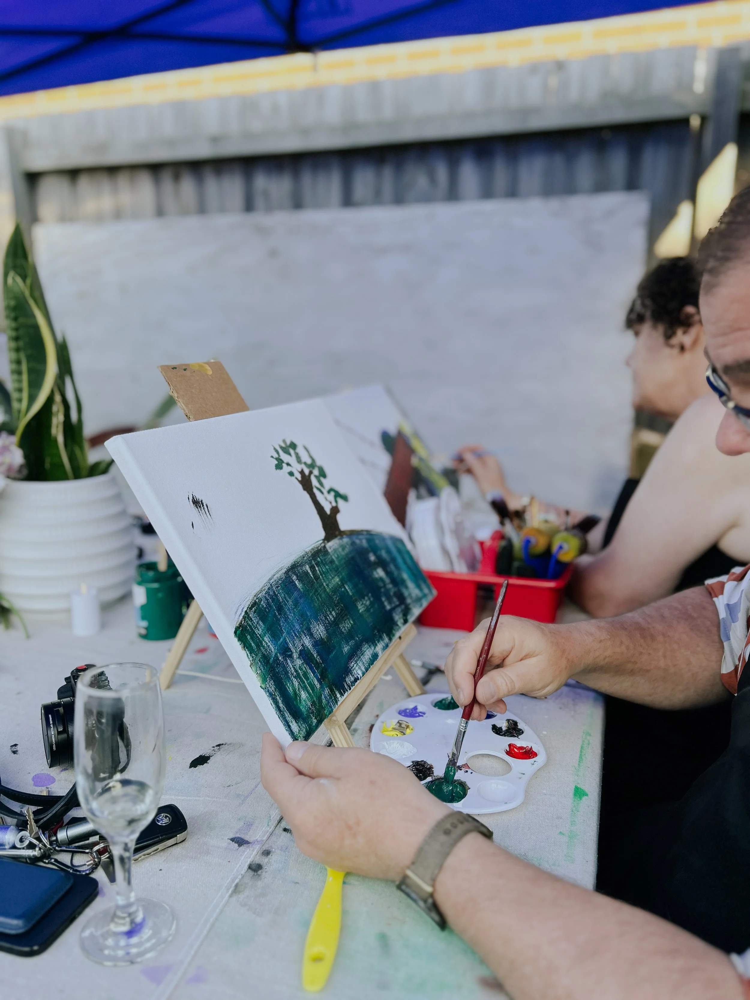 Person painting a landscape scene on canvas with a tree on a hill, using a palette of multiple colors, seated at a table outdoors with art supplies and a glass of champagne nearby.