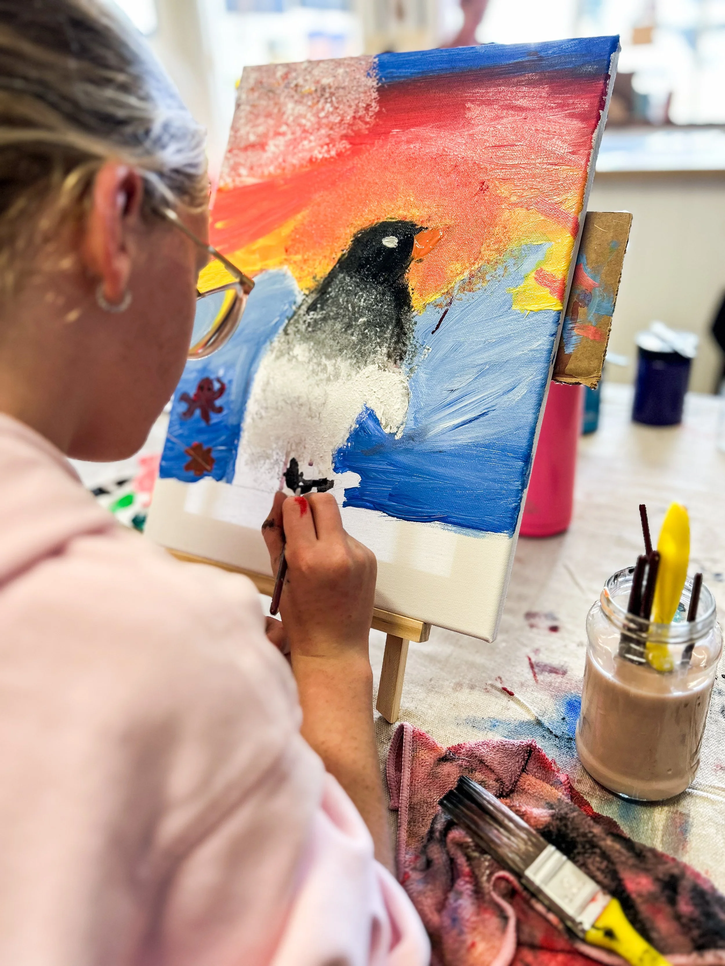 Person painting a penguin on a canvas with a colorful sunset background.