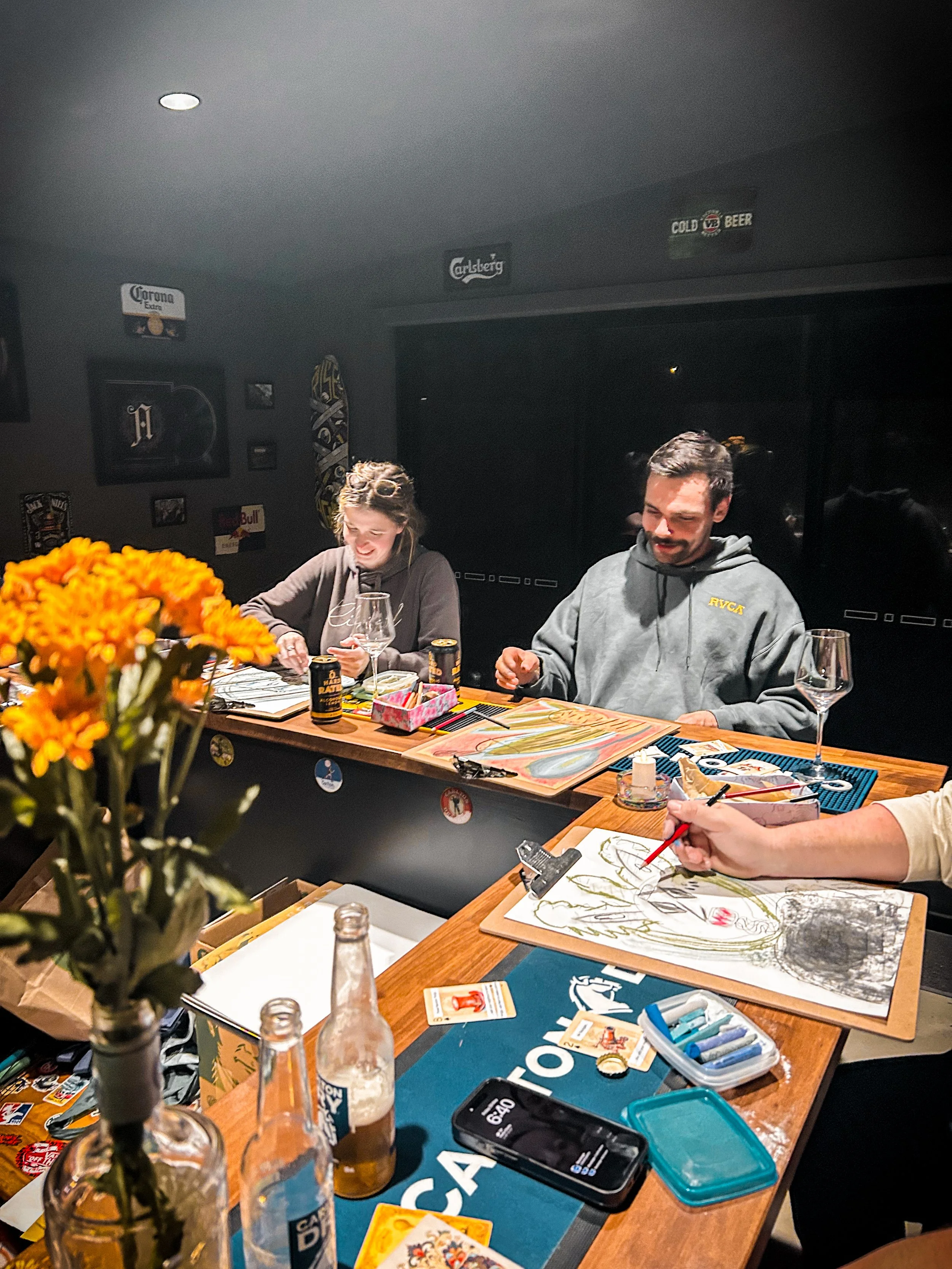 A group of people sitting around a table engaged in art and card games, with art supplies, drinks, and decorations like flowers.