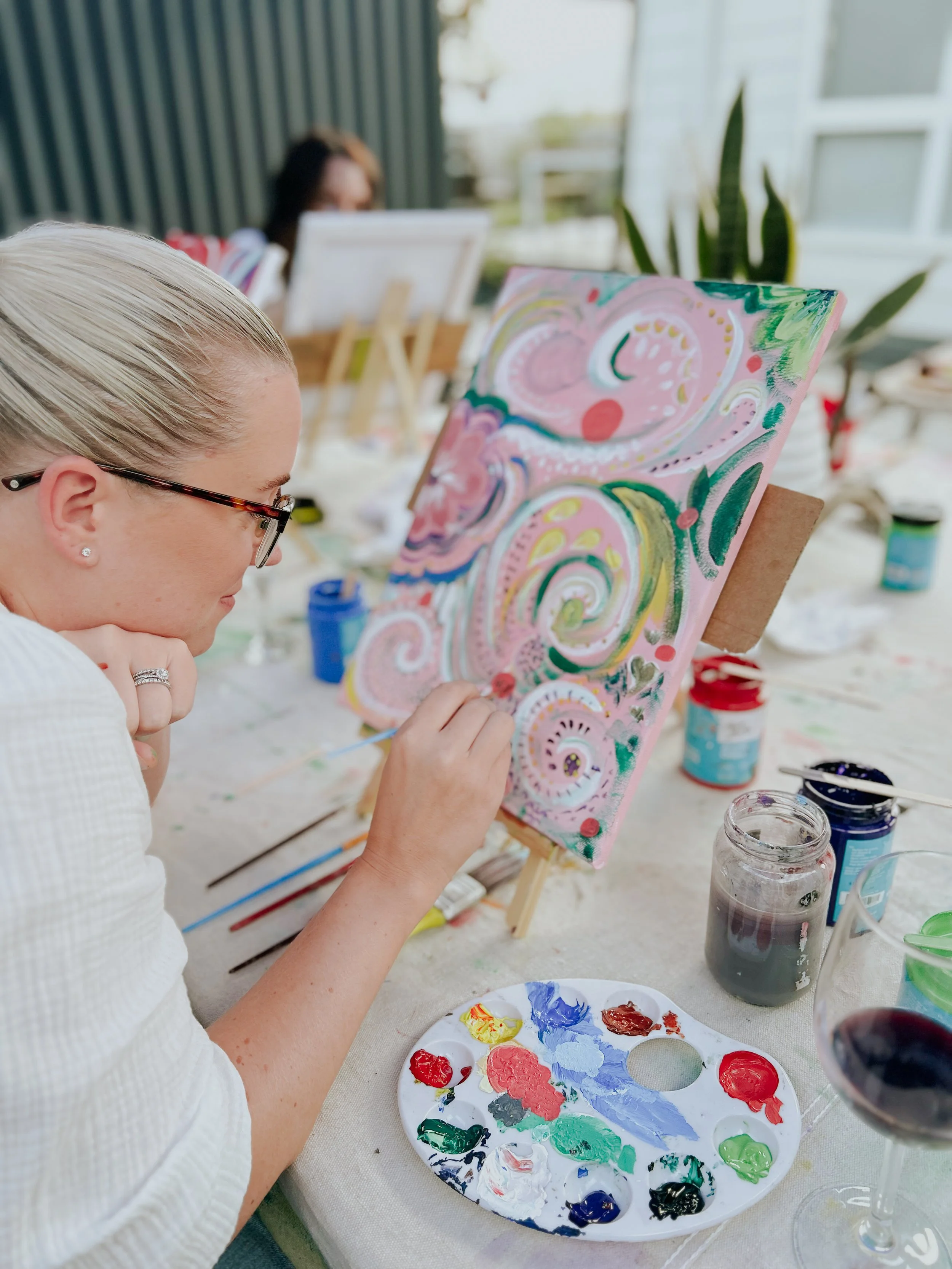 Woman painting a colorful, swirling abstract acrylic artwork on canvas, seated at a table with paint supplies, wine glass, and jars of paint in an outdoor setting.