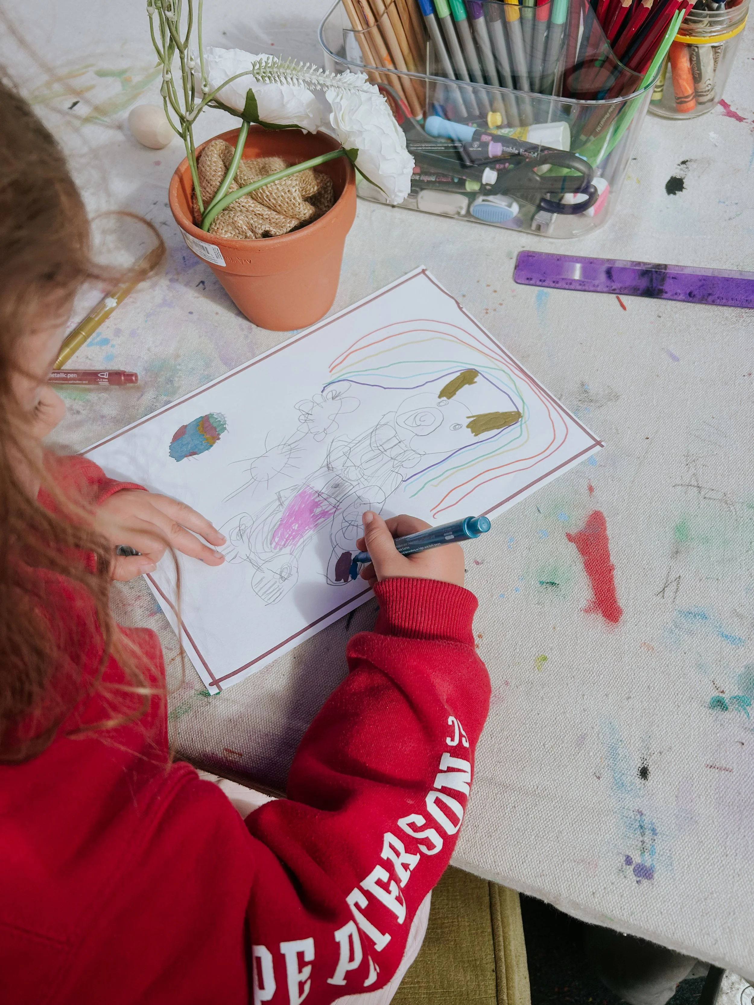 Child in a red sweatshirt drawing on paper with colored markers, with art supplies and a plant on a paint-splattered table.