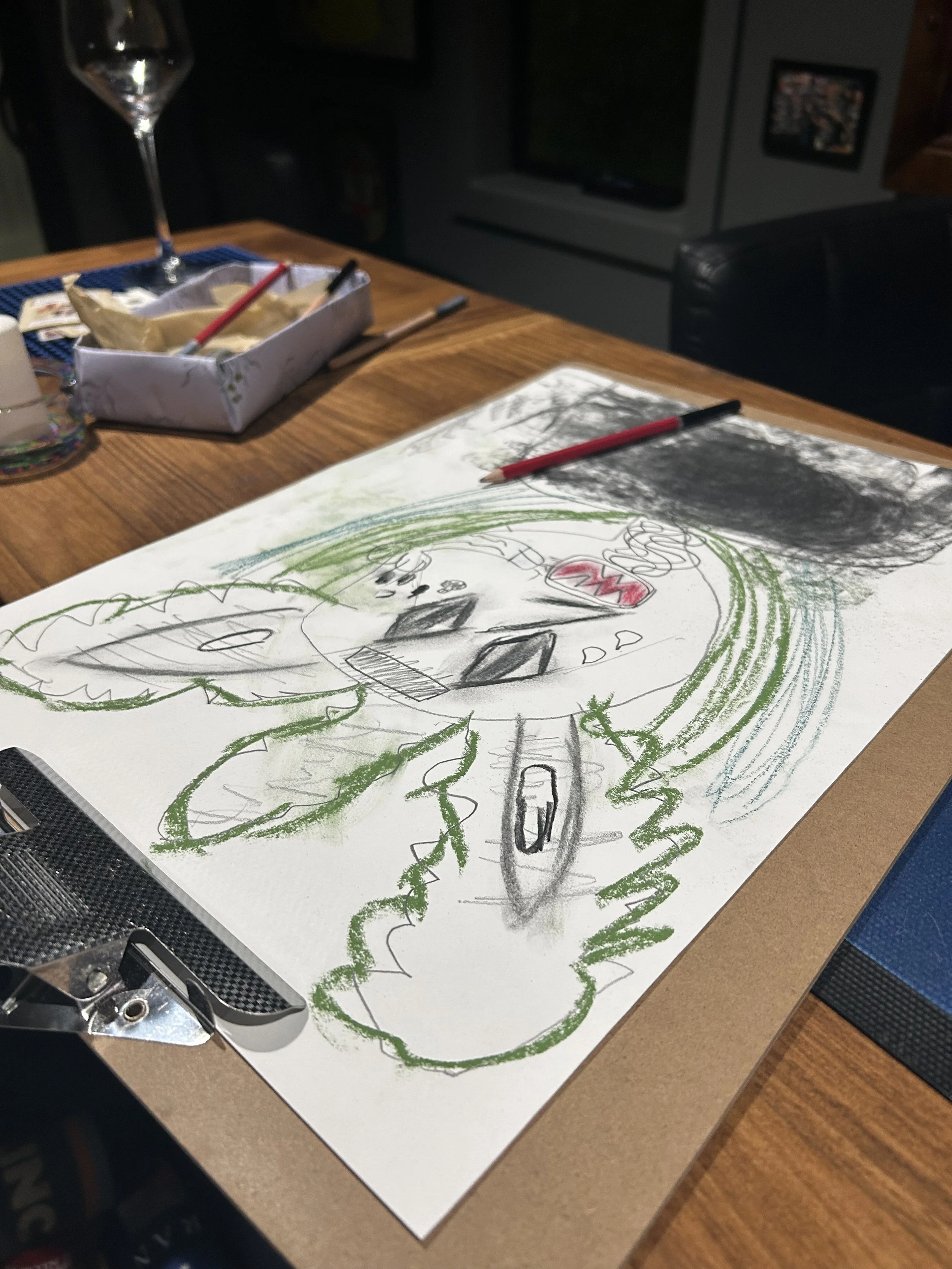 A drawing on a white sheet of paper depicts a cartoonish train with smiling face, colorful wheels, and a green cloud of smoke, lying on a wooden table. There are art supplies around, including colored pencils, a box with wrapping paper and an envelop