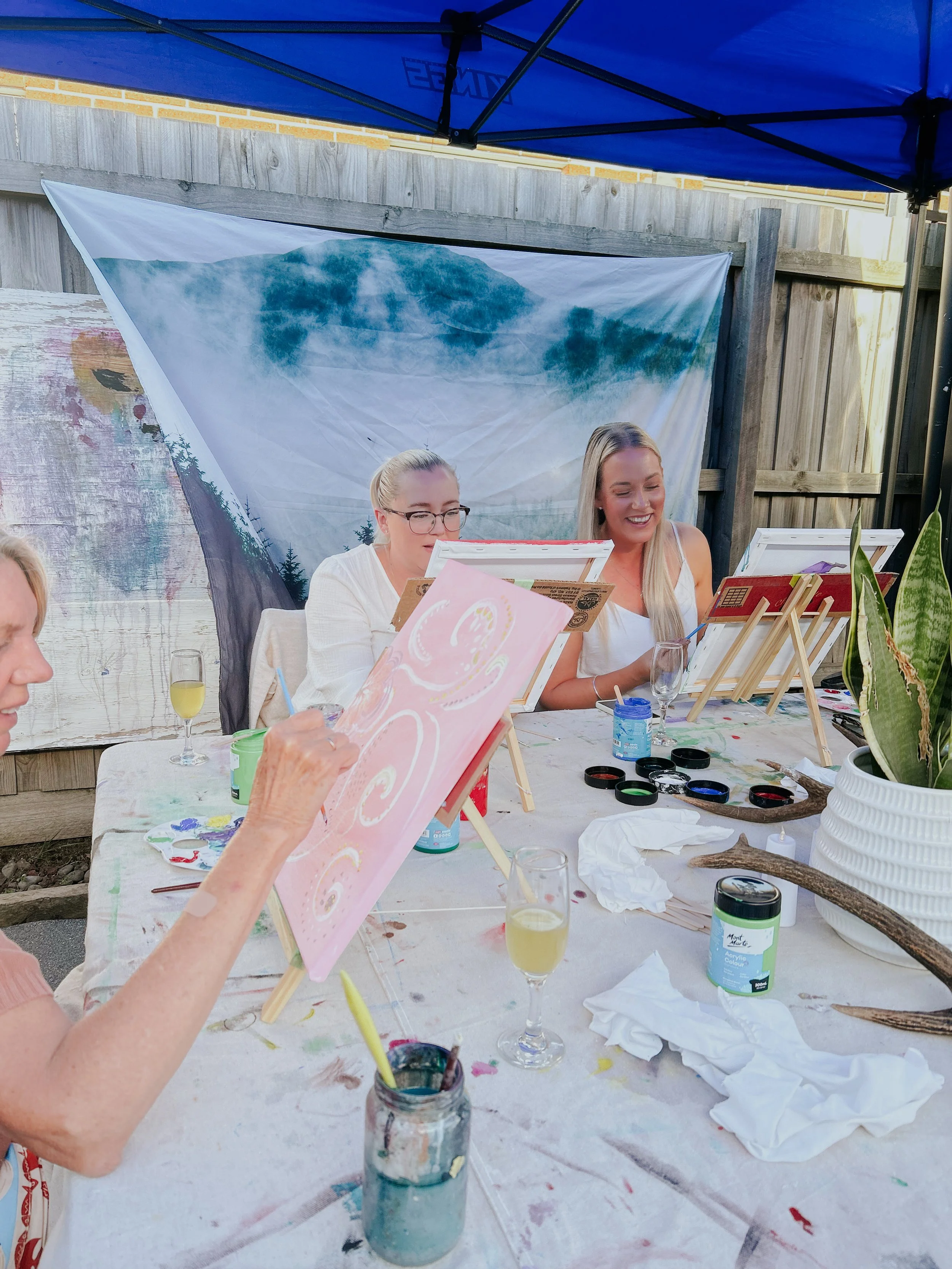 People participating in outdoor painting activity, seated at a table with painting supplies, drinks, and a scenic backdrop.