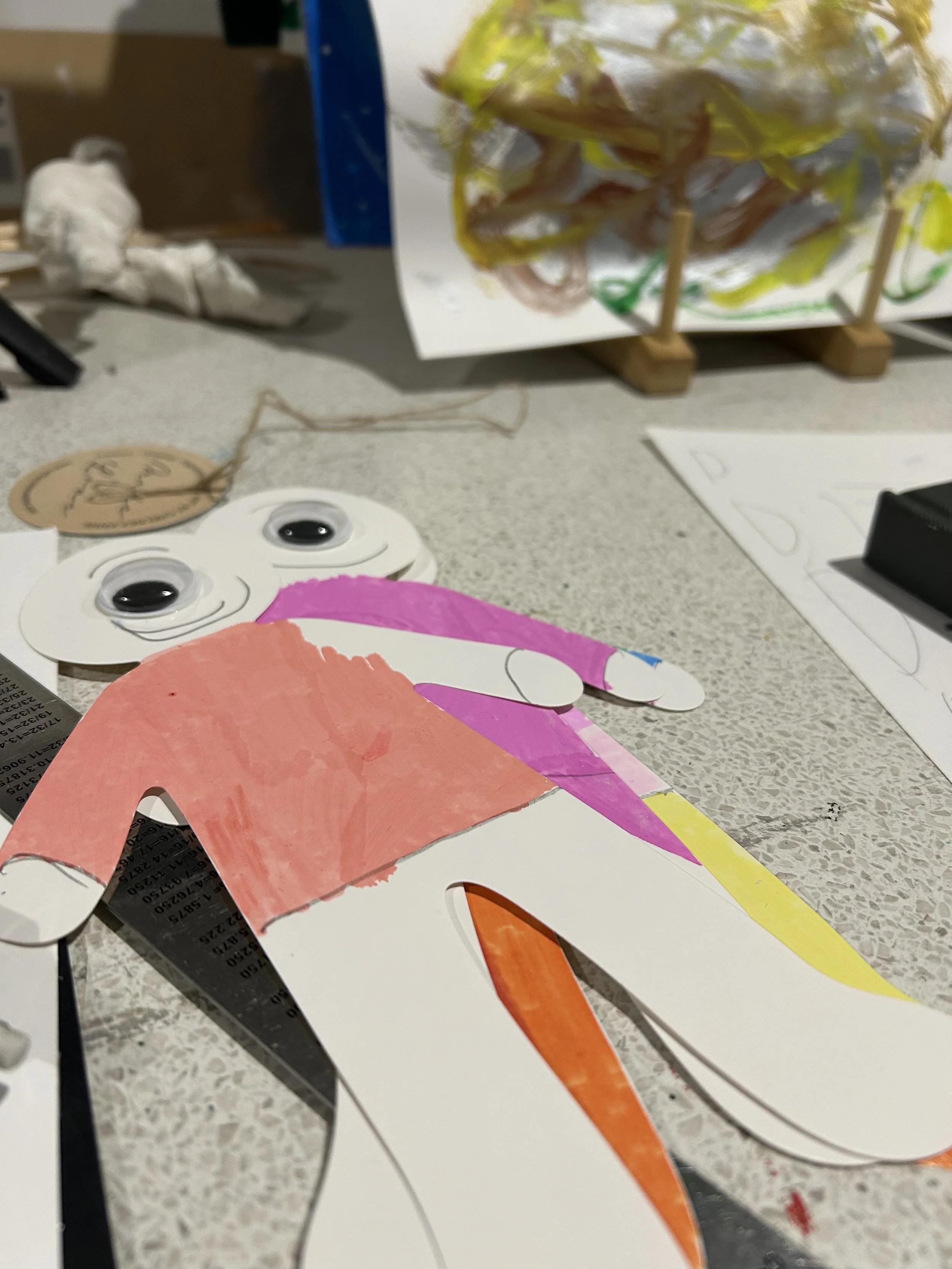 Colourful paper craft of a cyclops with large googly eyes, positioned on a workspace with other craft materials and drawings.
