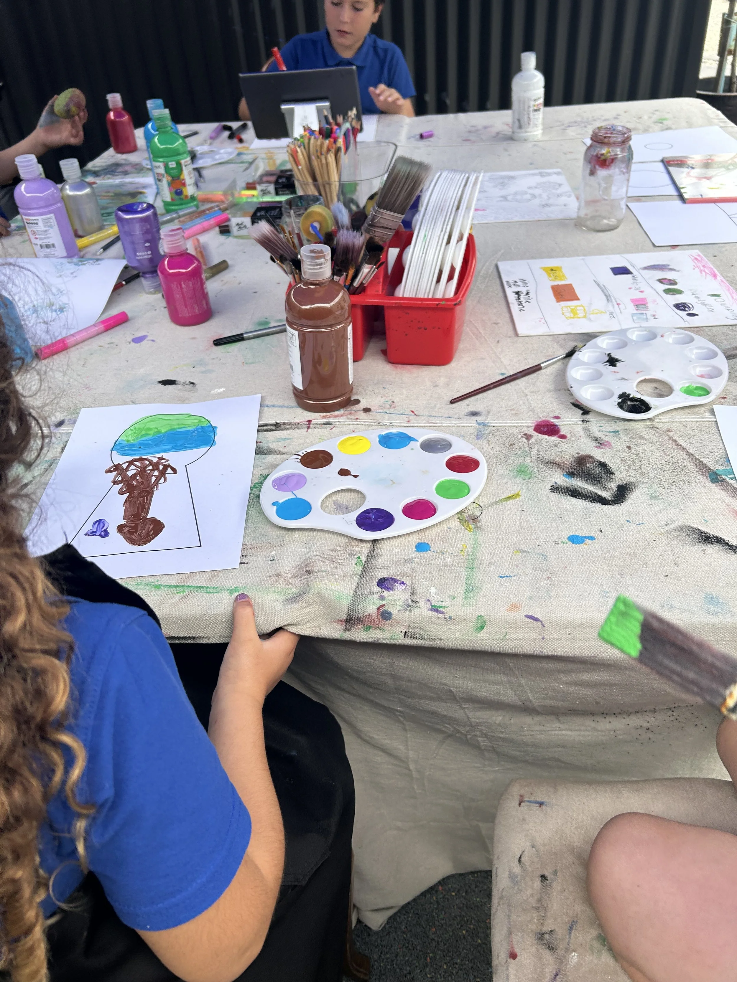 Children painting and drawing at a messy table with paint palettes, brushes, and art supplies.