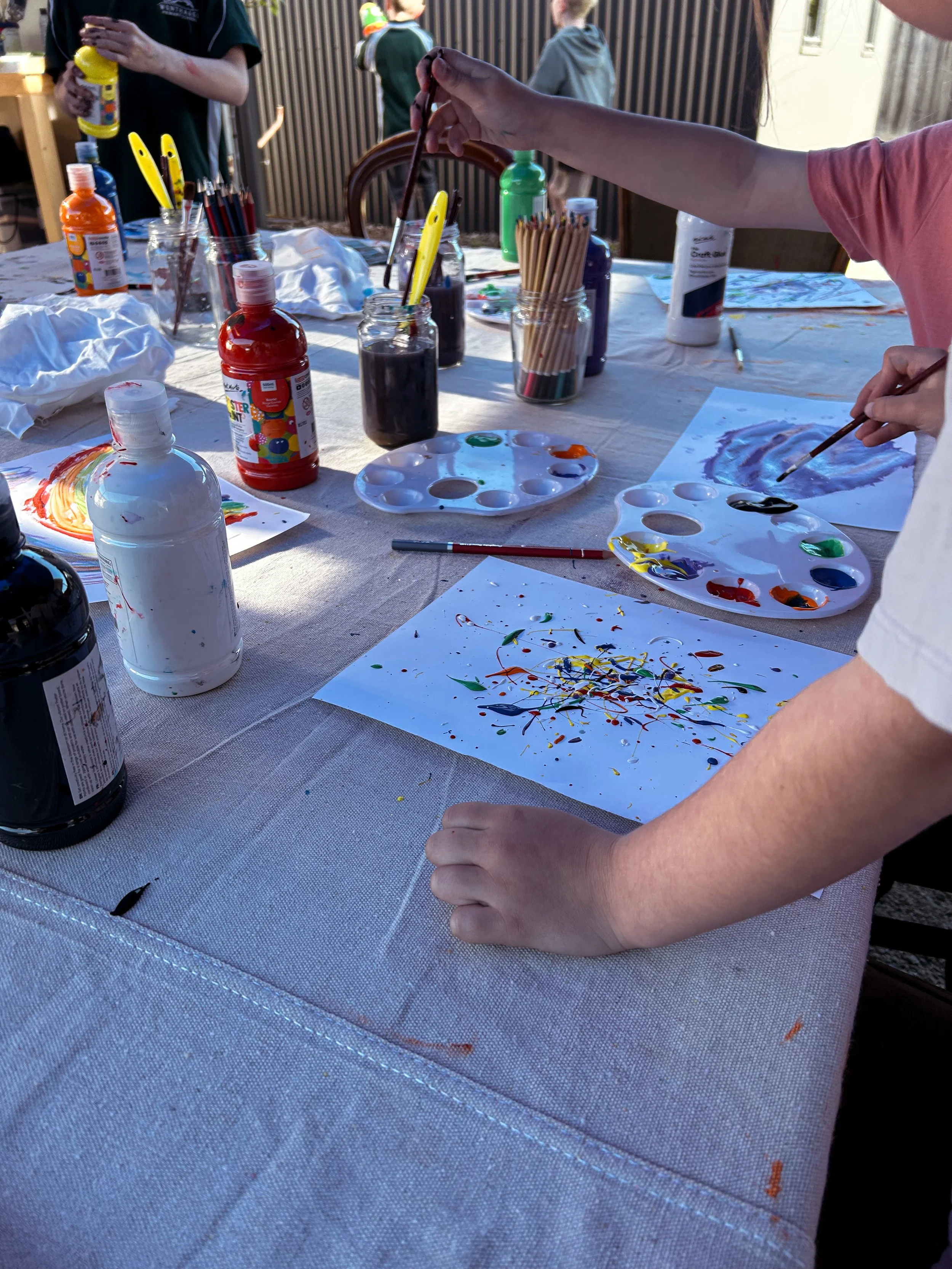 Children participating in a painting activity outdoors with colorful paints and paper on a table.