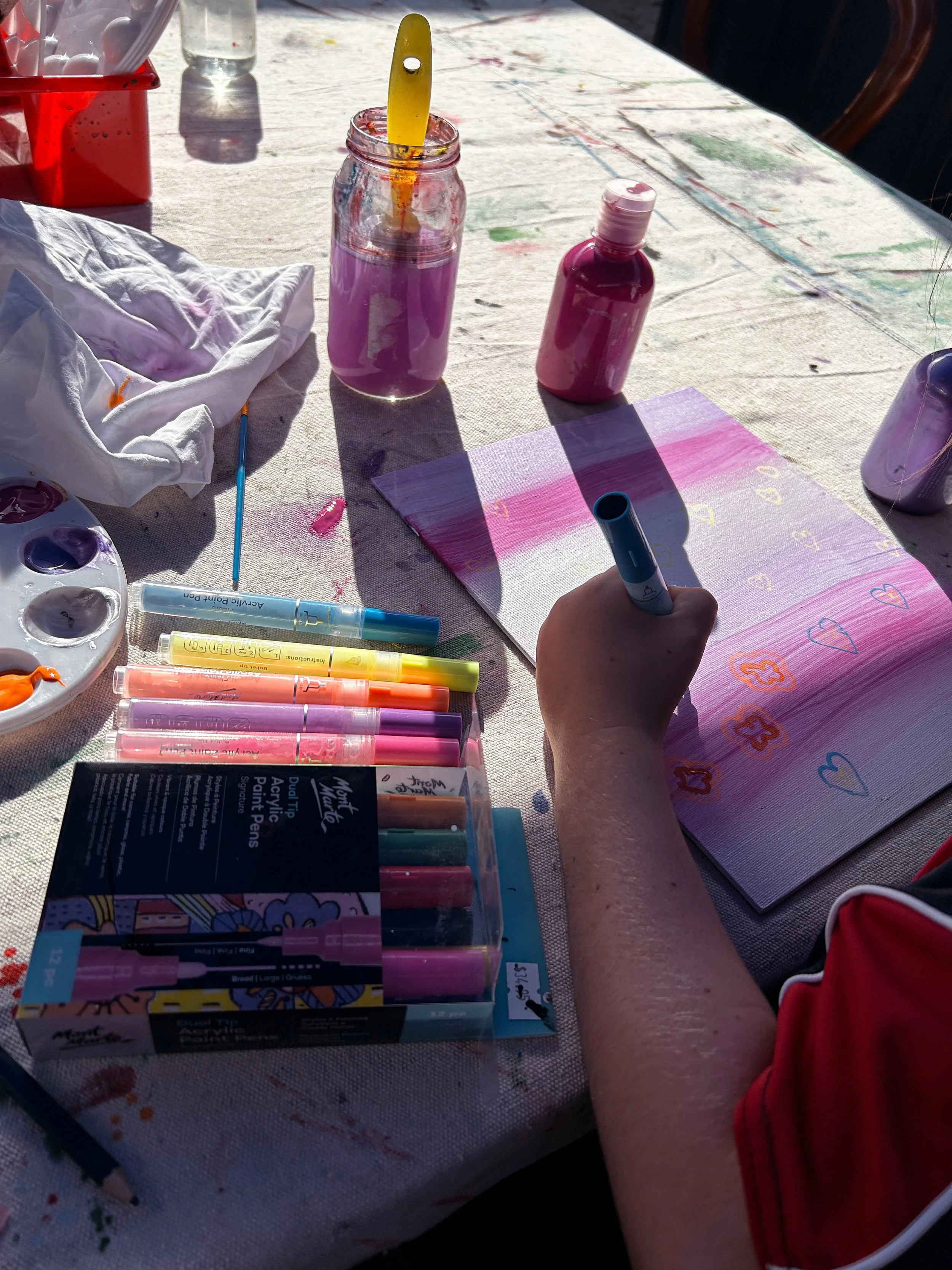 Person painting a colorful design with markers on paper at a table with paint supplies nearby.