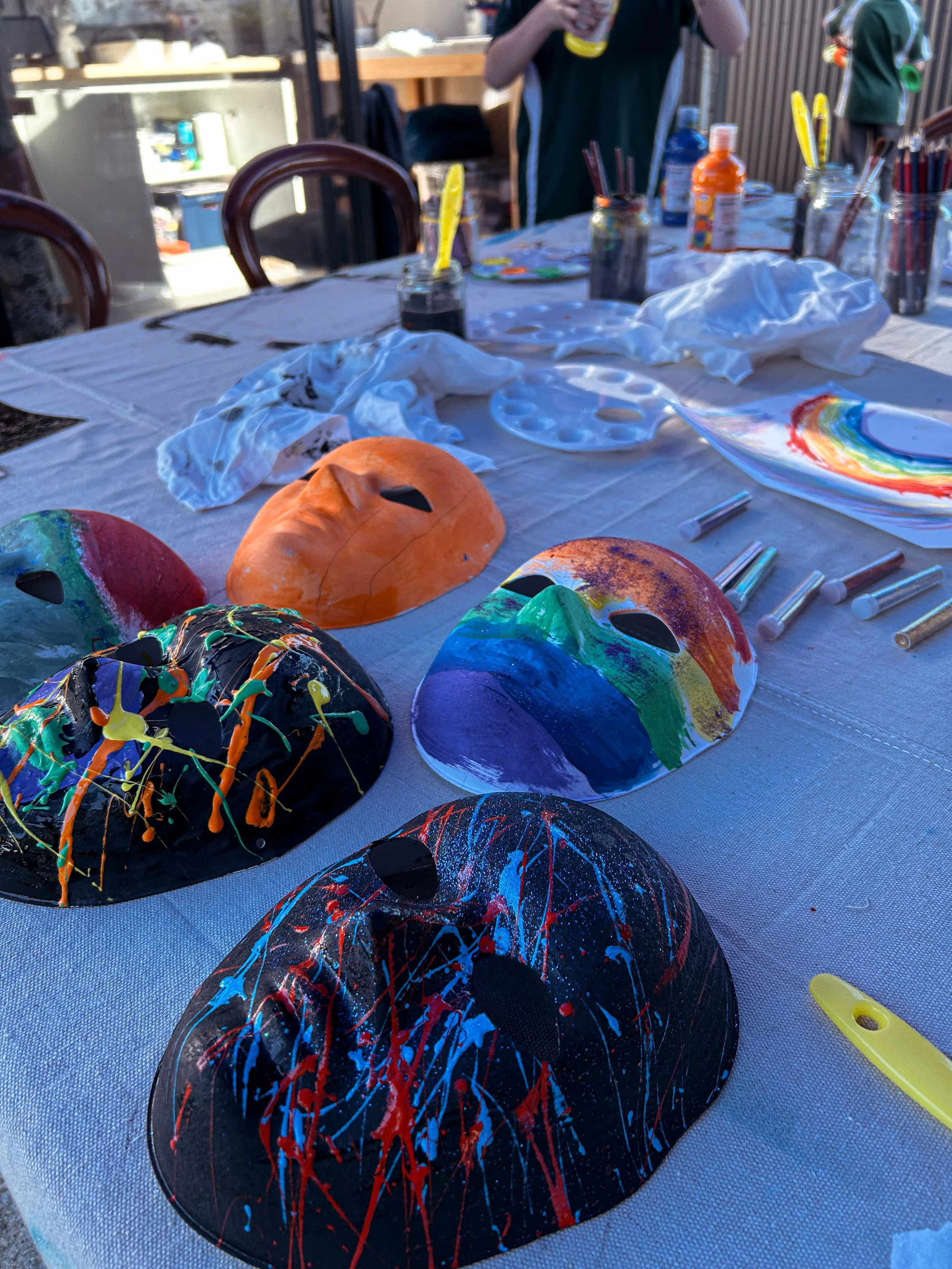 Colorful, painted masks on a table at an outdoor craft activity, with painting supplies and gloves around.