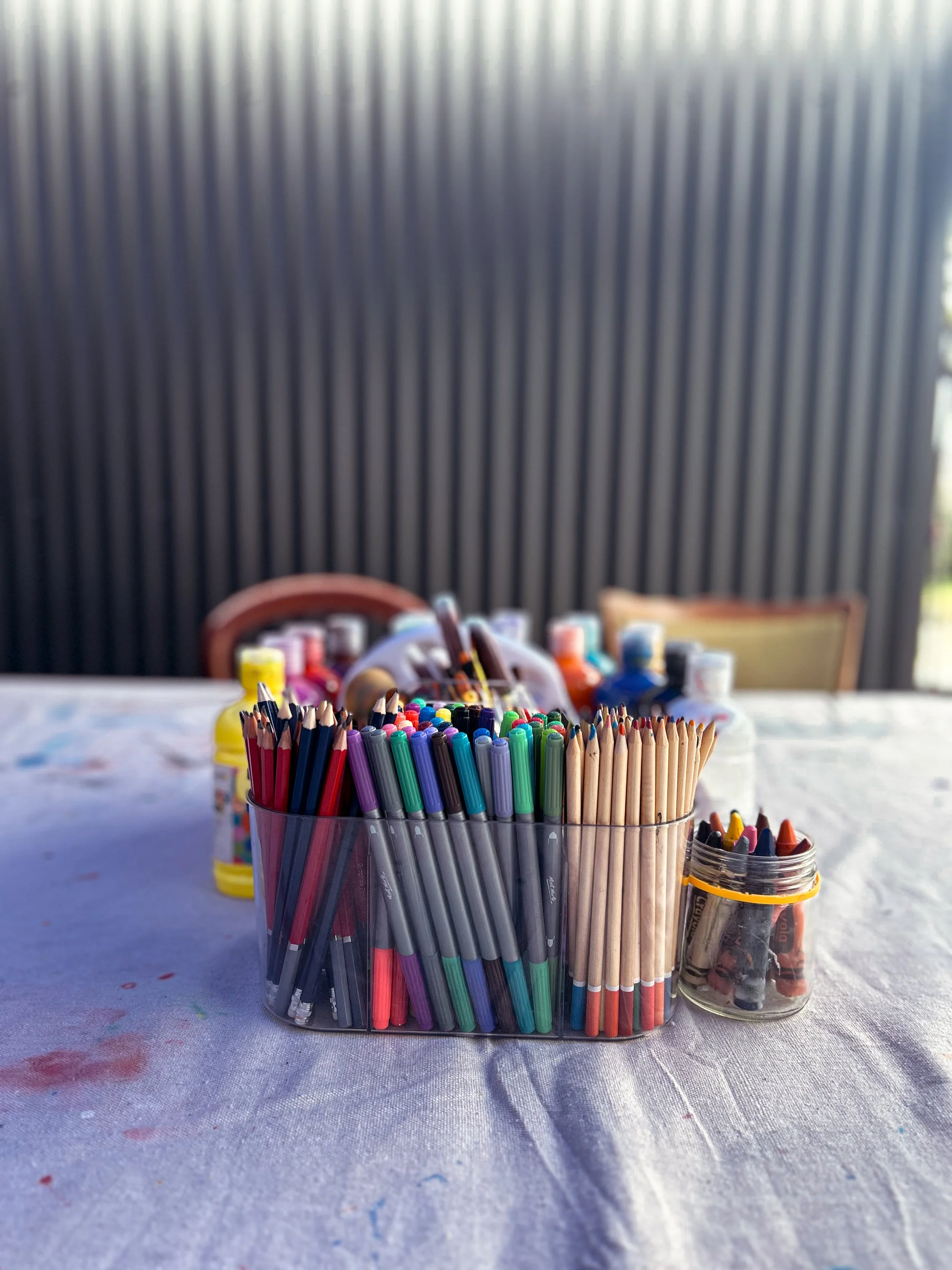 A container of colored pencils on a table with art supplies and chairs in the background.
