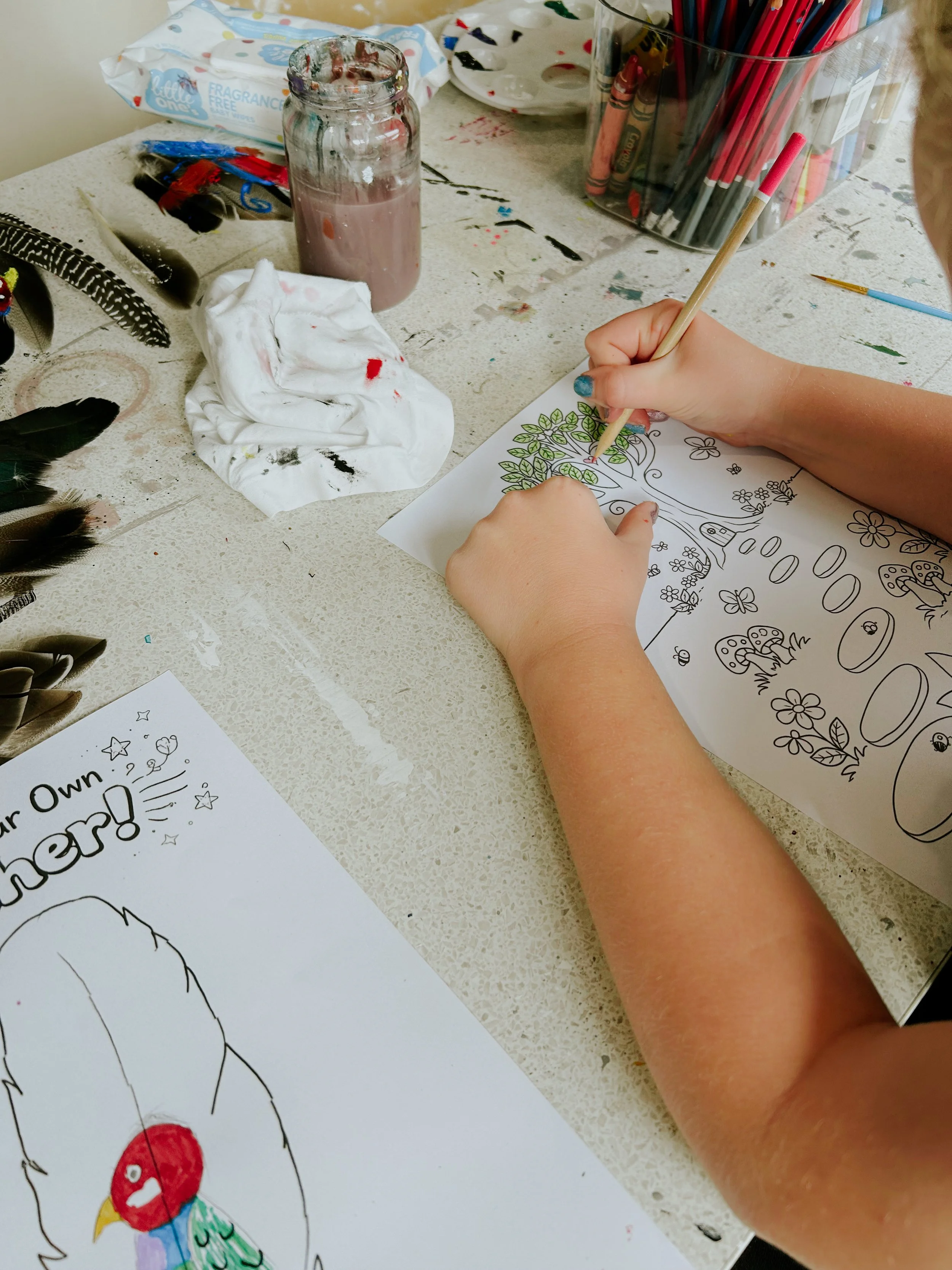 Child's hands coloring a drawing of a bird and a tree with a coloring page and markers on a table.