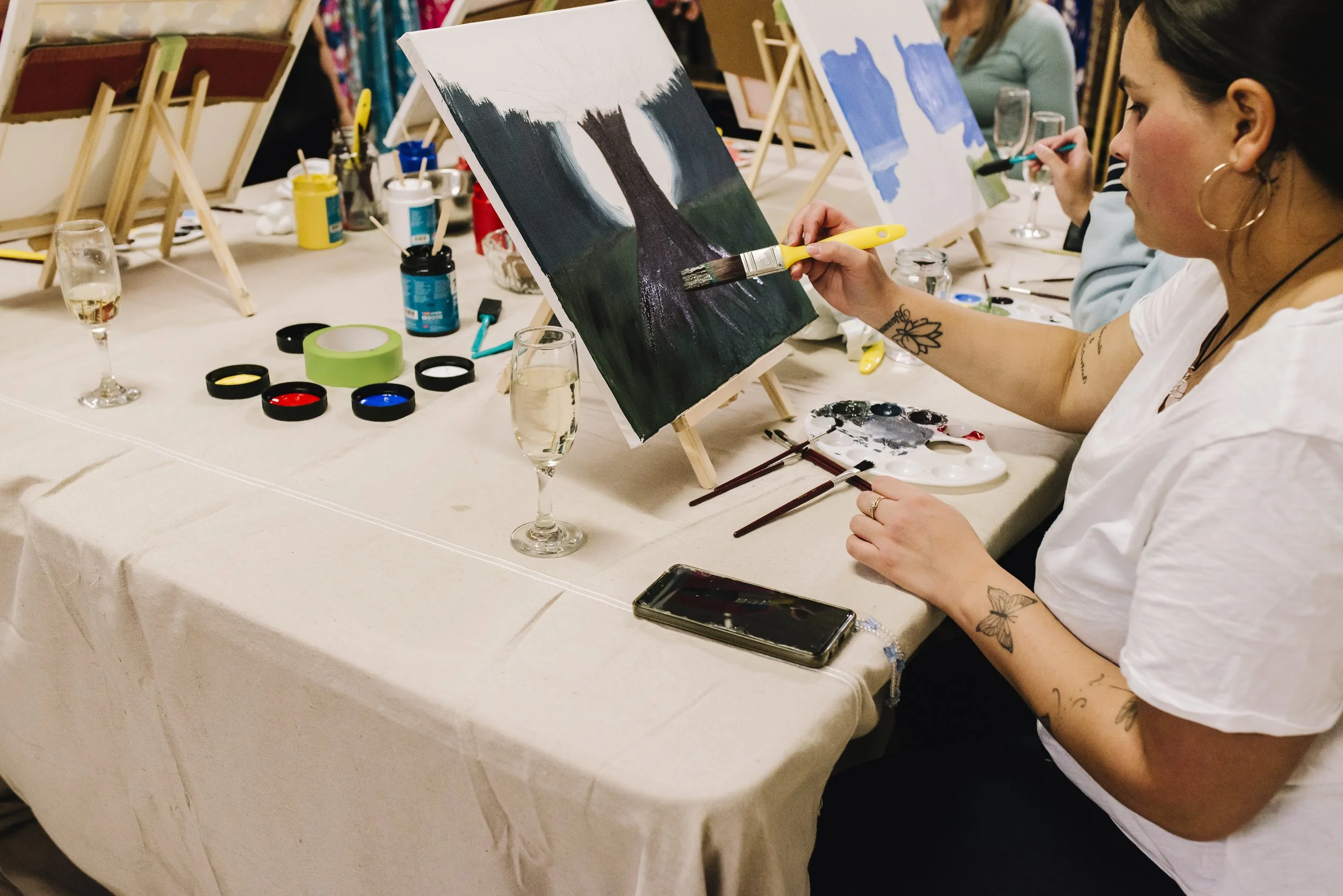 A woman painting a landscape on a canvas at an art class, with bingo-style paints and brushes on the table, and glasses of champagne nearby.