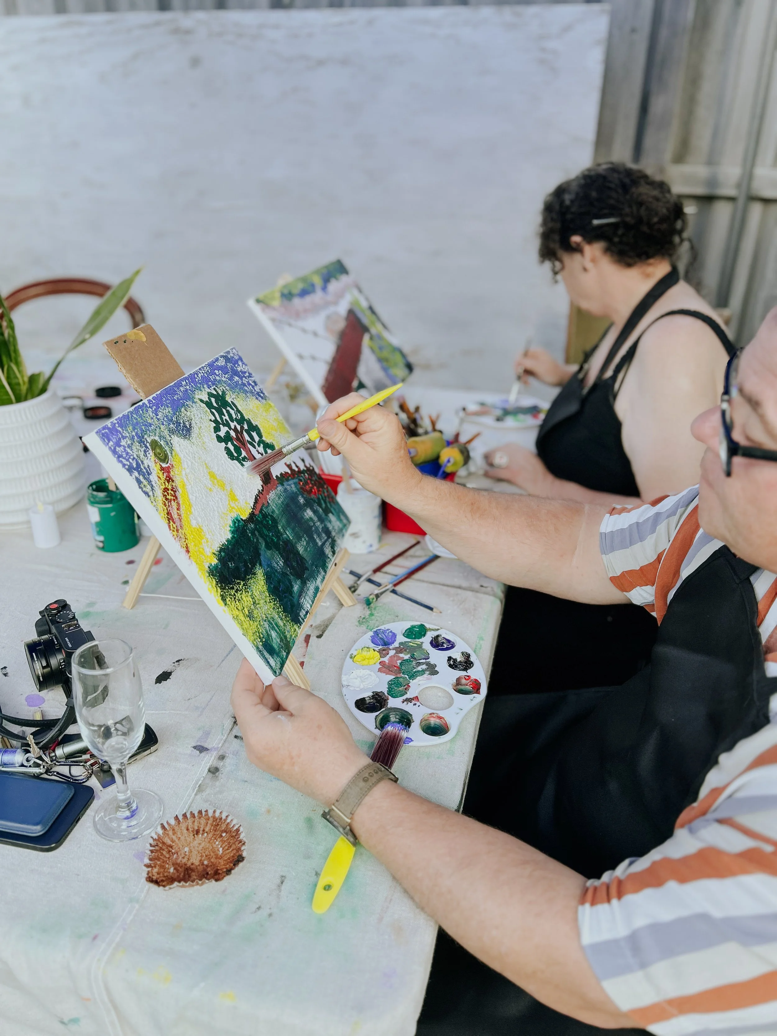 Two people painting colorful landscape scenes on small canvases with brushes at a table outdoors.