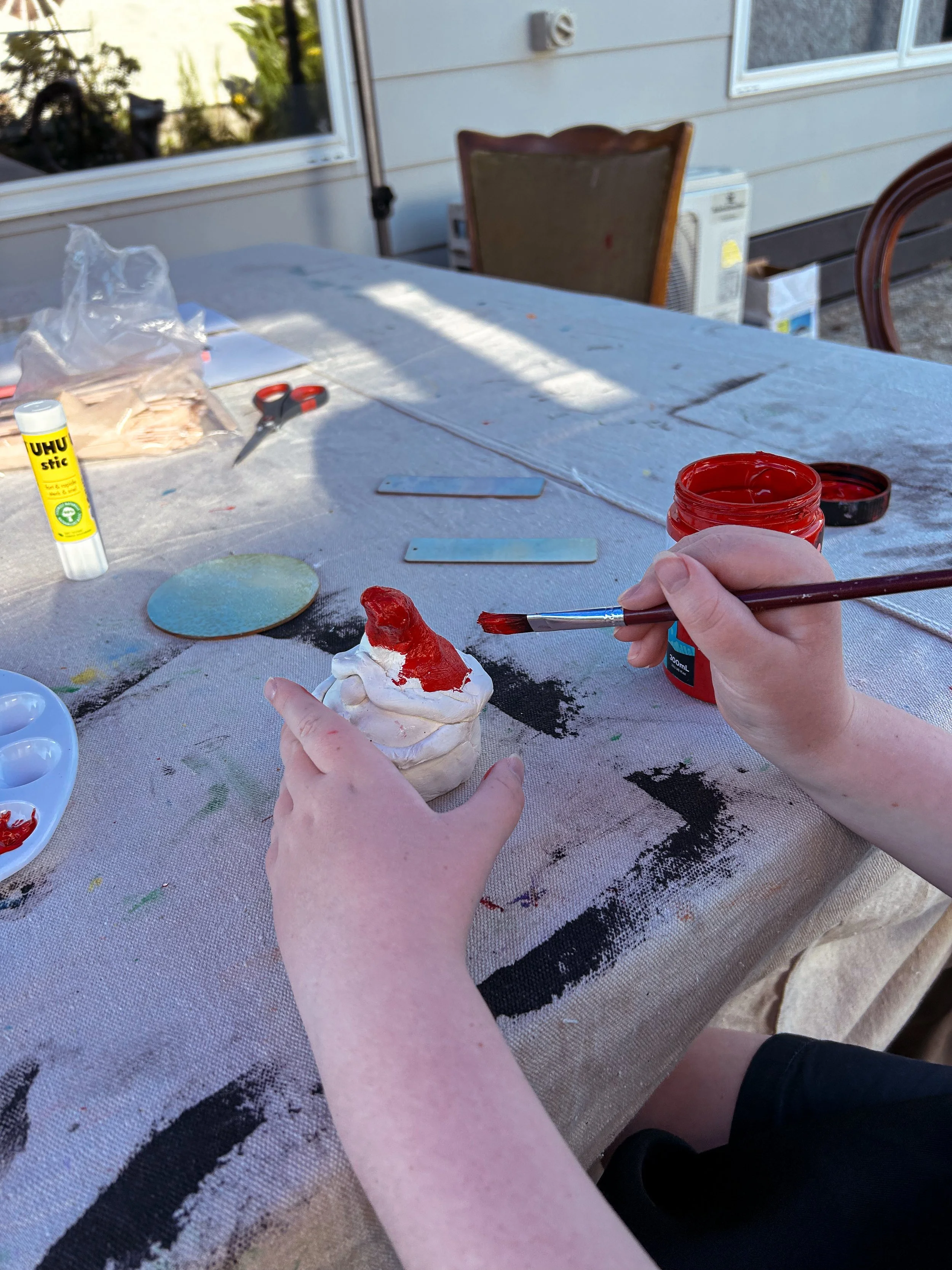 Person painting a Santa hat on a ceramic Christmas ornament outdoor table with paintbrush and red paint, with crafting supplies and tools around.