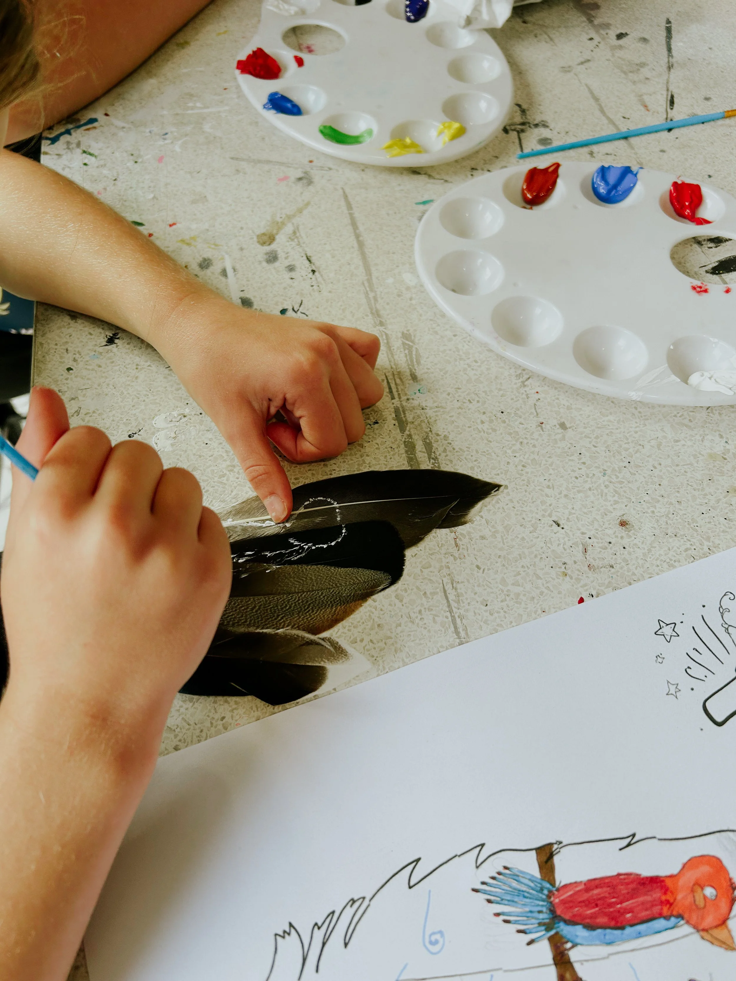 A child's hand and part of an arm are shown reaching towards a drawing of a colorful parrot on white paper. Two paint palettes with various colors and a paintbrush are visible on a work surface nearby.