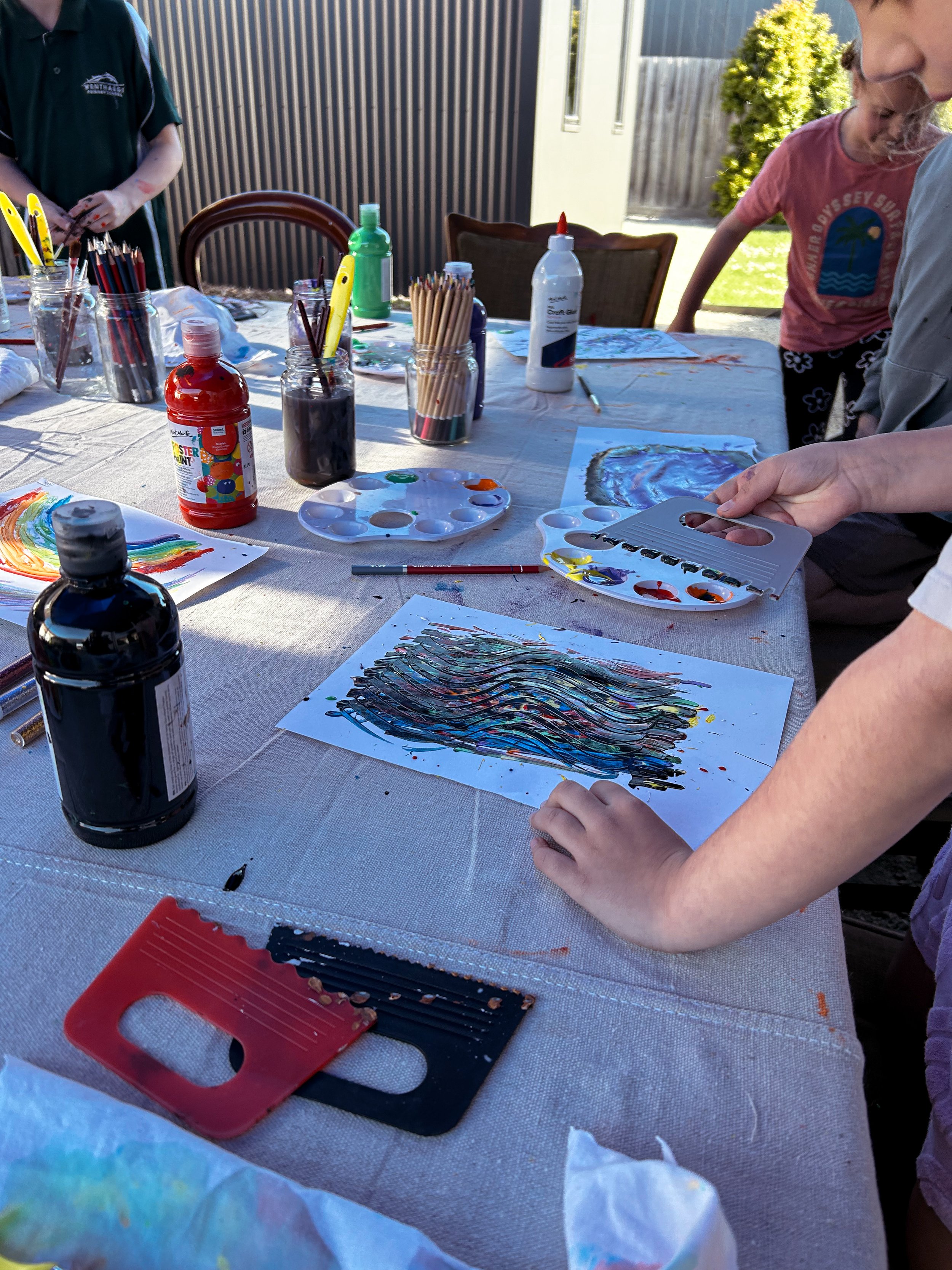 Children and adults painting and creating artwork outdoors at a table covered with art supplies, including paints, brushes, and paper.