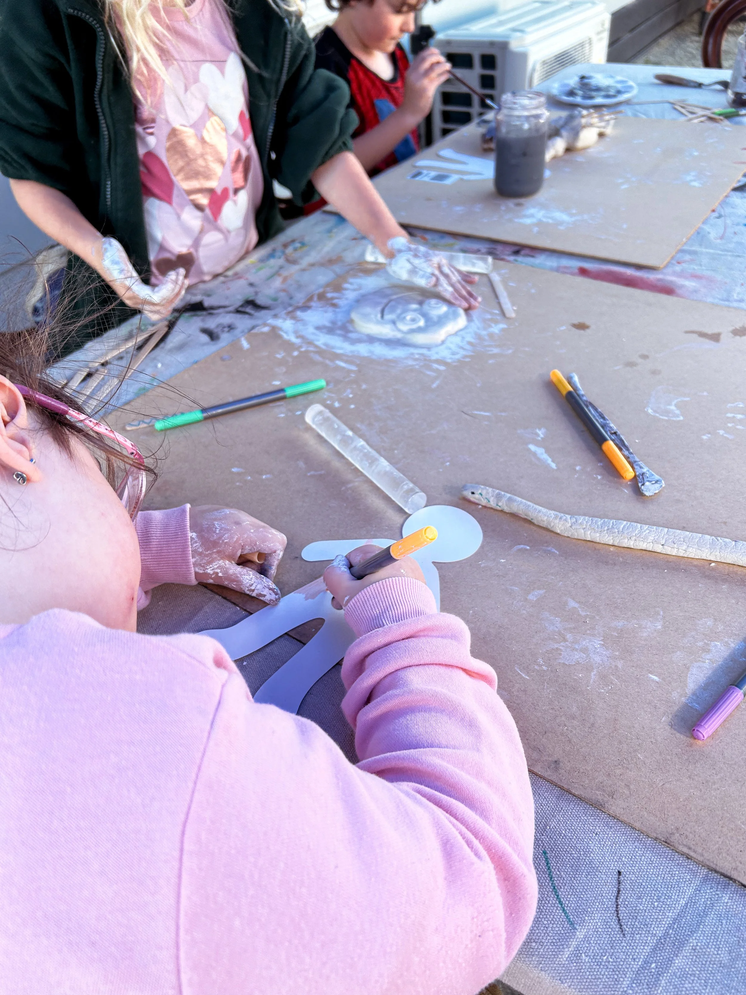 Children creating art on a large table outdoors, with art supplies scattered around, including markers, scissors, and a paper cutout.