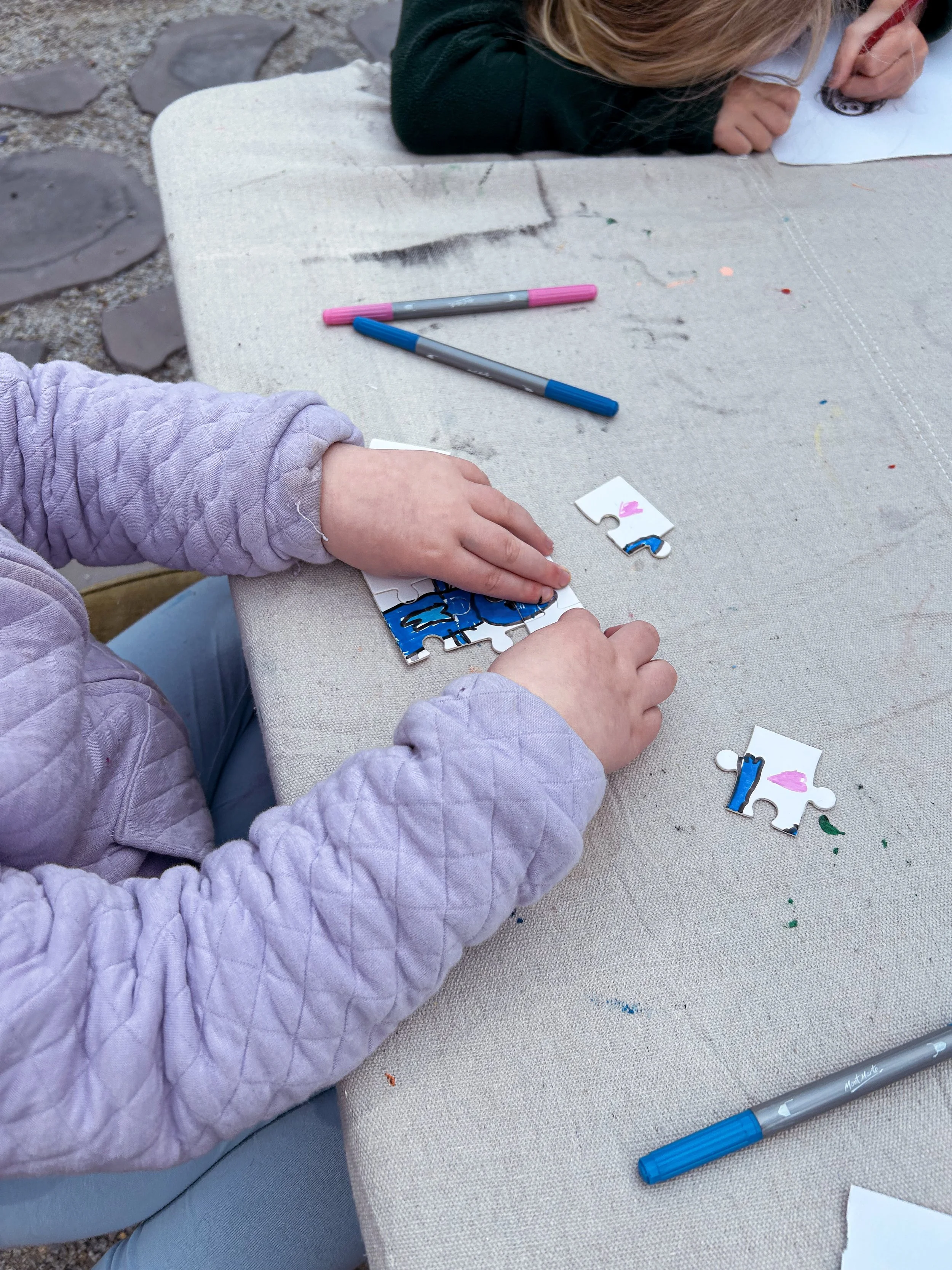 Child assembling a puzzle at a table outdoors. The puzzle features a blue cartoon character. Two markers, one pink and one blue, are on the table.