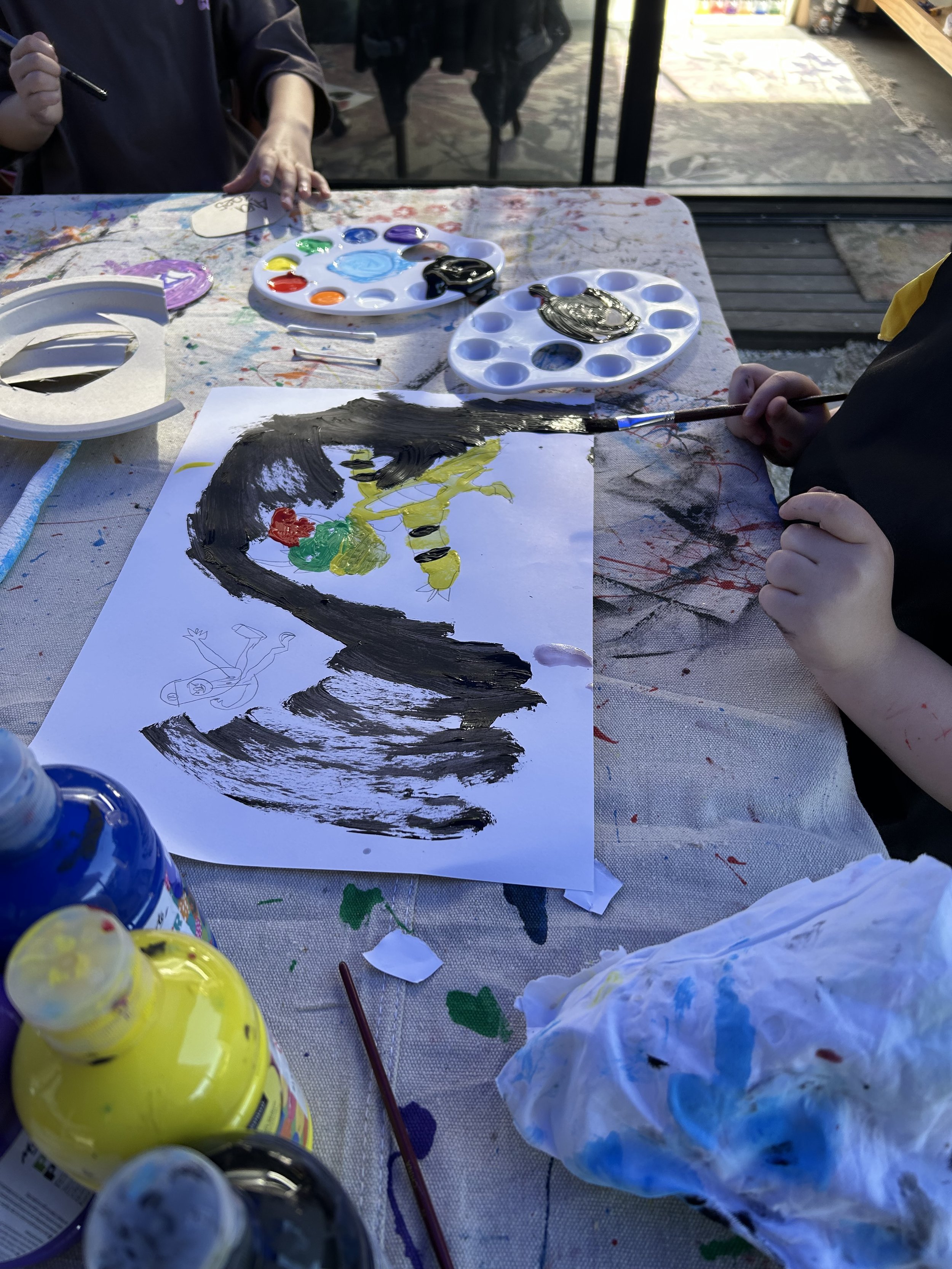 Child painting a colorful picture with black, yellow, green, and red paint on paper, surrounded by paint bottles, palettes, and art supplies on a messy table outdoors.