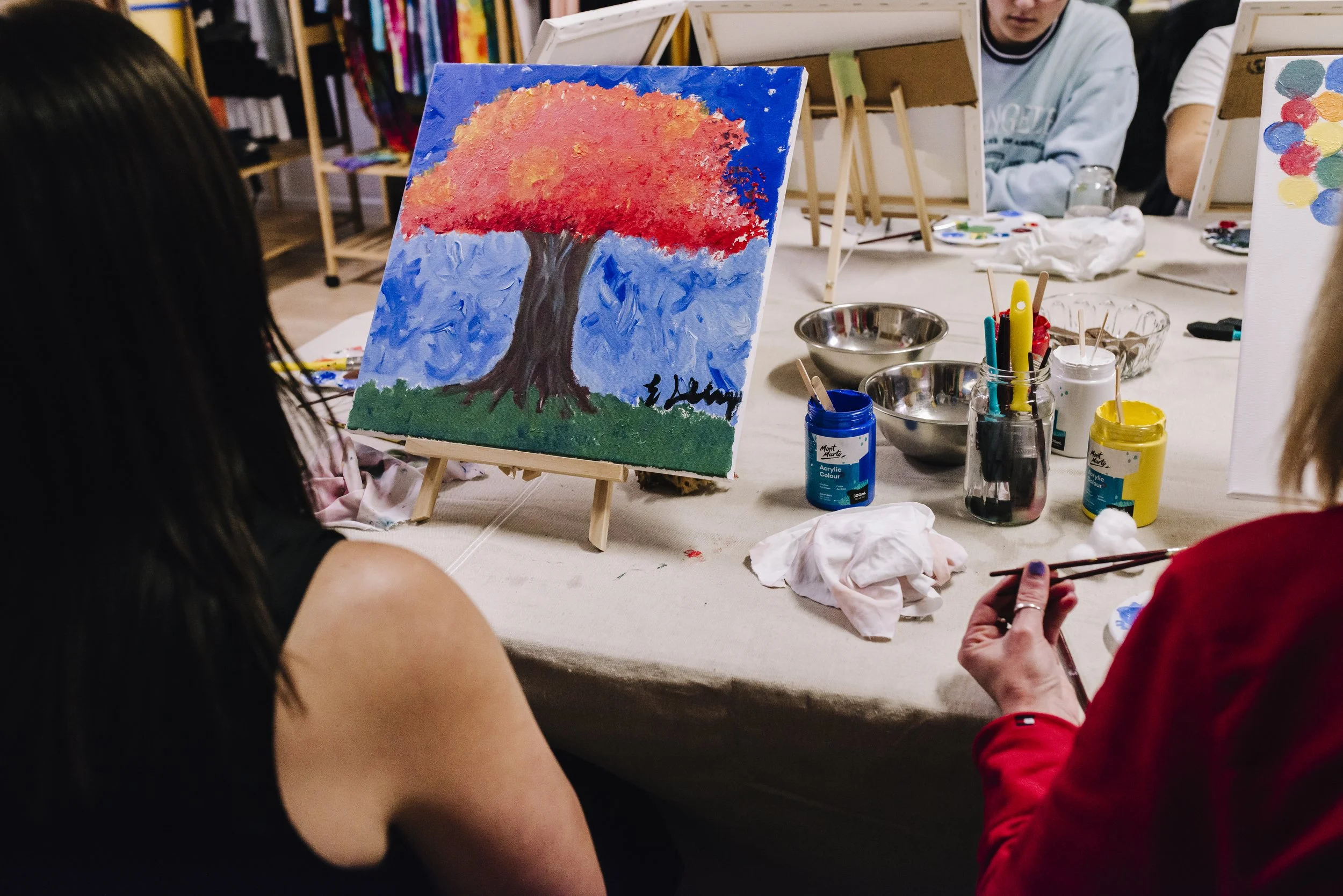A group of people painting on canvas at a table, with a finished painting of a colorful autumn tree on an easel in the center.