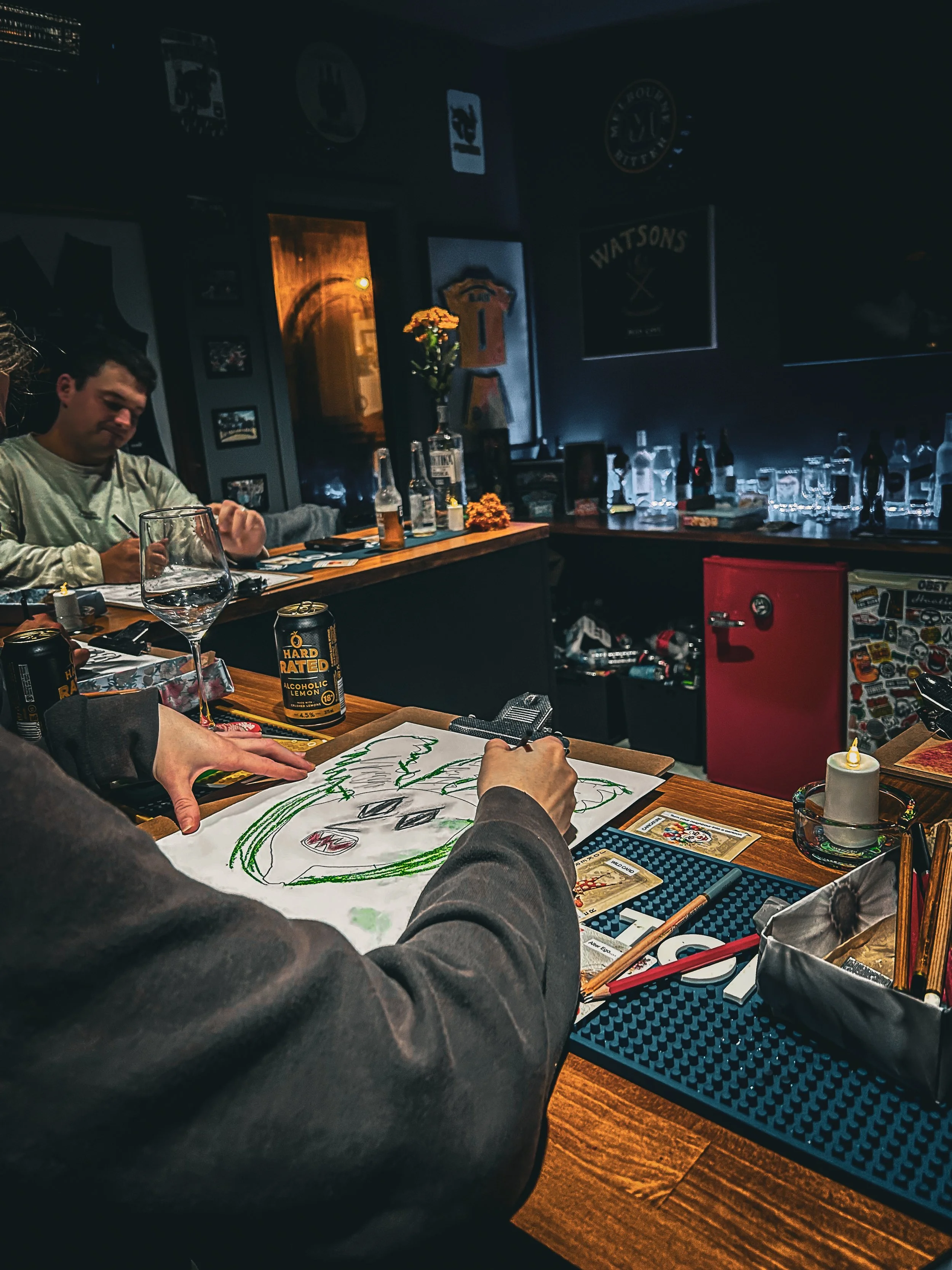 A group of people playing a card game at a bar. One person is drawing a large face with green hair on a piece of paper with coloring supplies. The bar has various bottles, glasses, and a Watson's poster on the wall.
