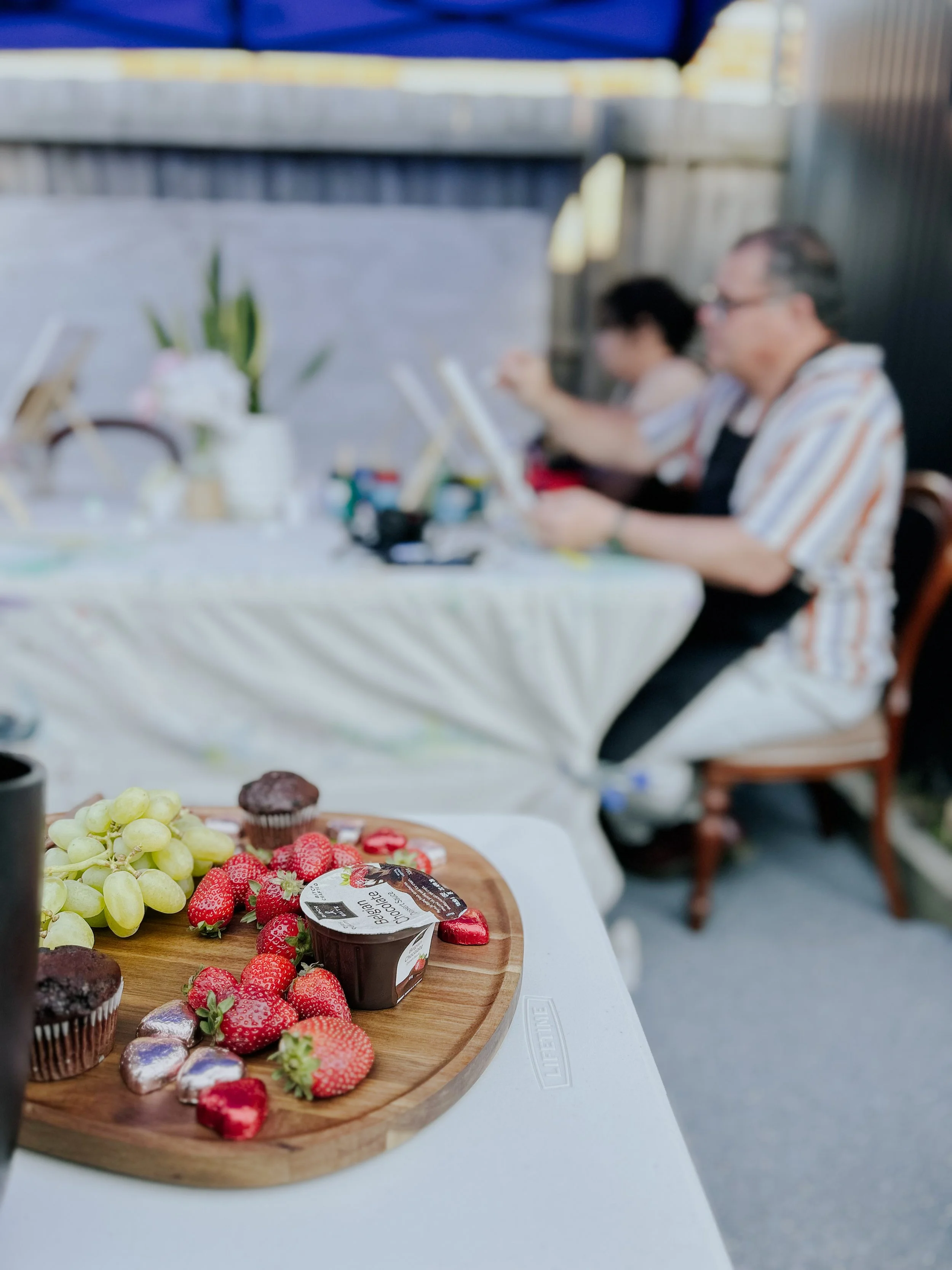 Fruits, strawberries, grapes, and small cupcakes on a wooden tray on a white table, with people sitting at a table in the background.