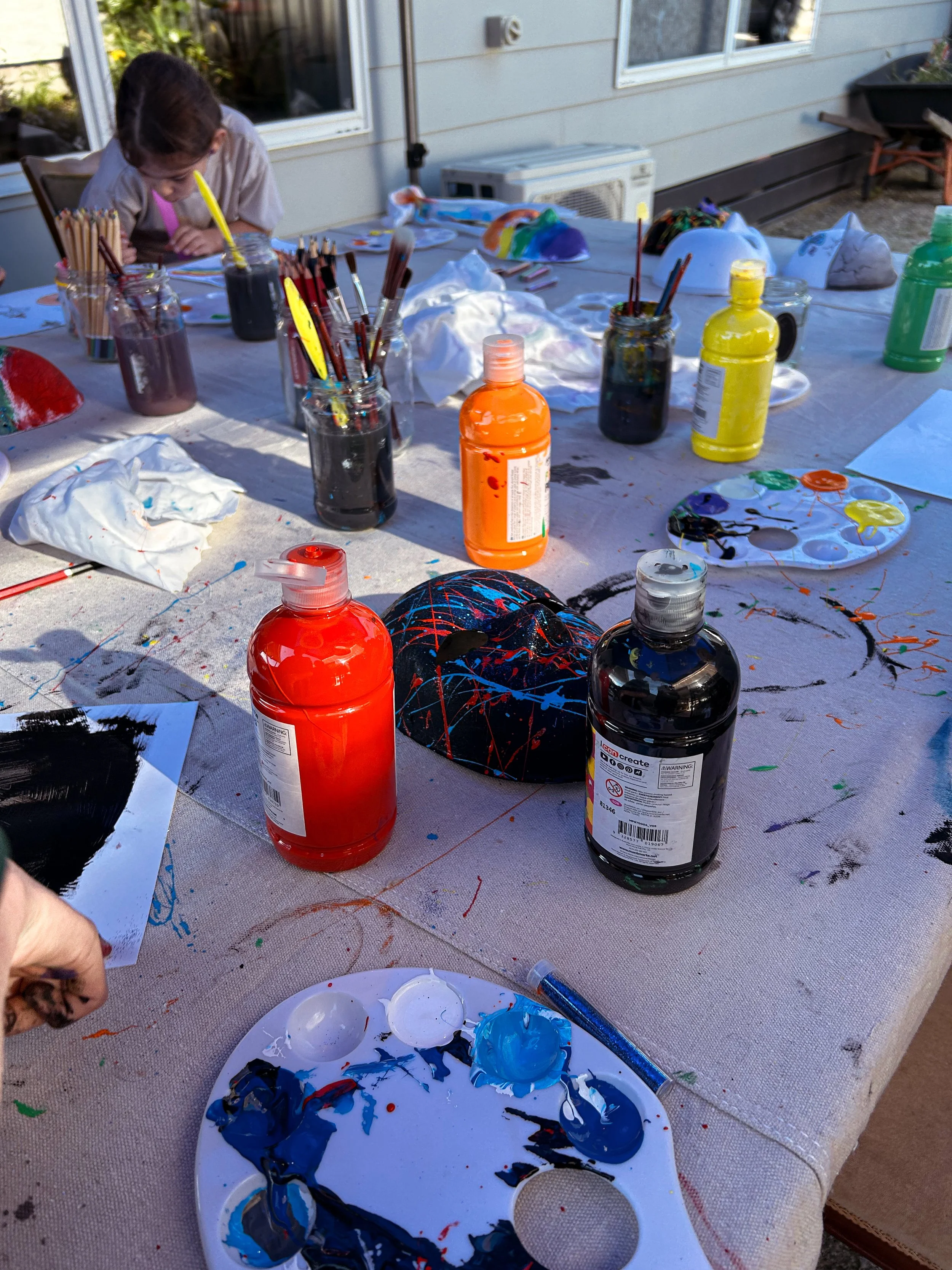 A table covered with painting supplies such as bottles of paint, paintbrushes, paints, a palette with mixed colors, and a person in the background working on an art project outdoors.