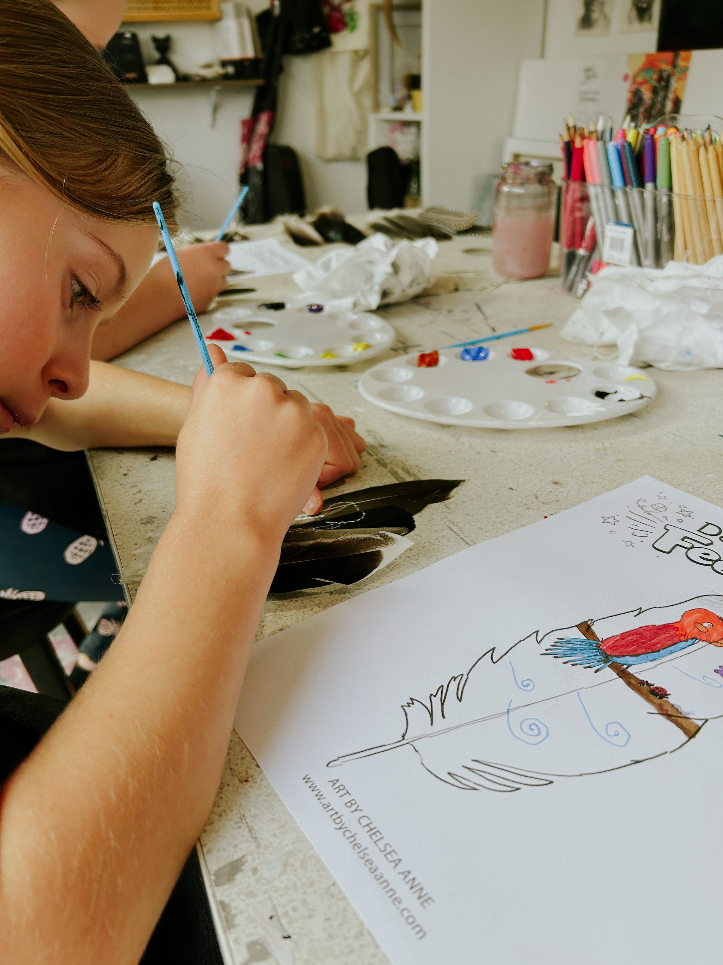 A child painting with a small brush at a table. Art supplies, including paint palettes, colored pencils, and paper with a bird drawing, are visible on the table.