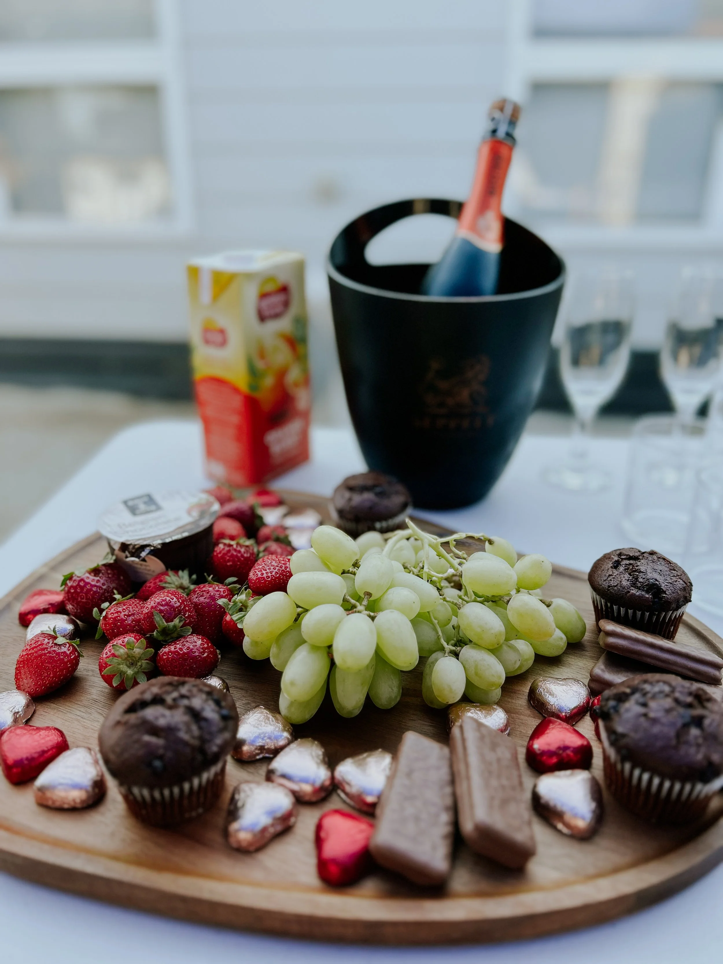 A wooden serving tray with strawberries, green grapes, chocolate muffins, heart-shaped chocolates, cookies, and a small container of chocolate spread. In the background, there are glasses, a carton of juice, and a champagne bottle in an ice bucket.