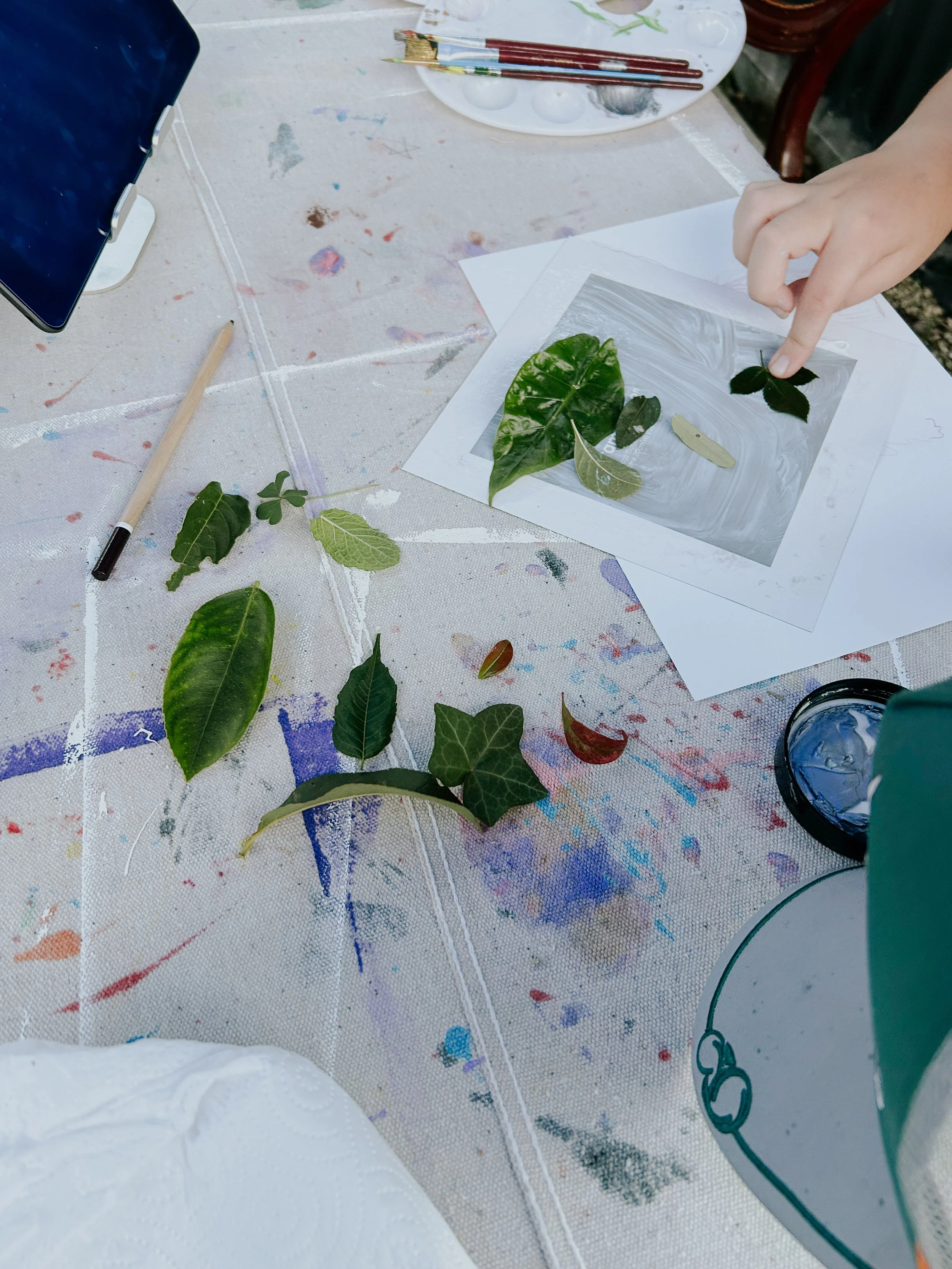 A person is pointing at a clear sheet with pressed green and brown leaves on it, placed on a white sheet of paper. The table has paint splatters and art supplies, including paintbrushes and a palette, nearby.