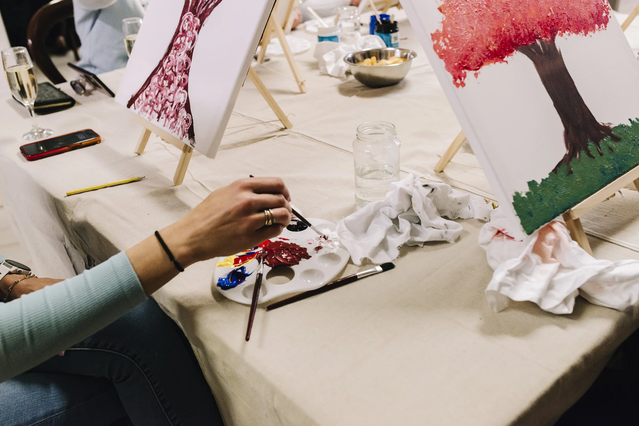 A person painting a colorful tree on a canvas. The table has painting supplies, including a palette with red, blue, and yellow paint, paintbrushes, rags, and a glass of water. Several finished or in-progress paintings are propped up on small easels.