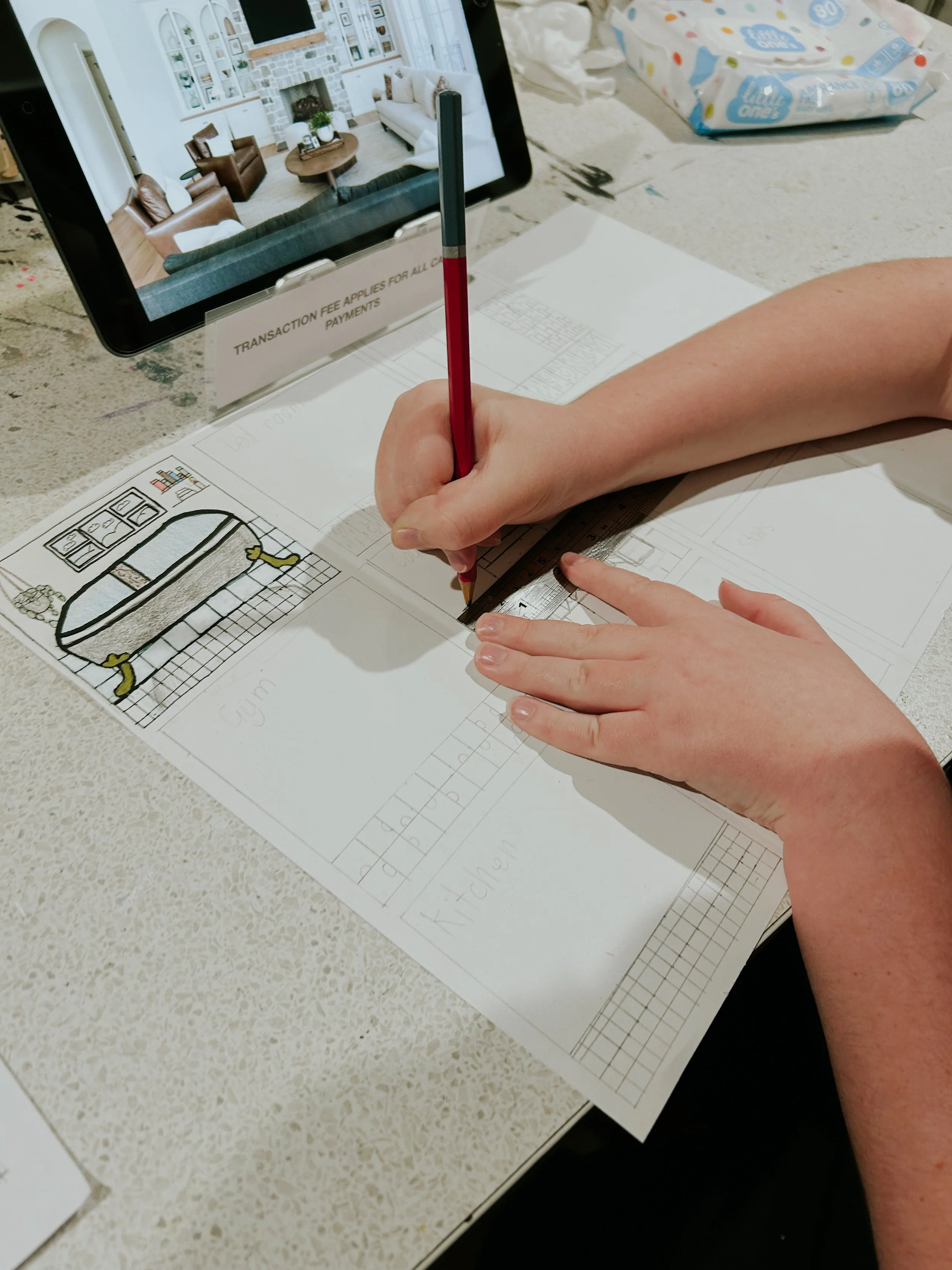 Person using a pencil and ruler to trace on a sheet of paper with a framed drawing of a sofa and armchair, on a white speckled tabletop, with a tablet displaying a living room interior and a pack of wipes or tissues nearby.